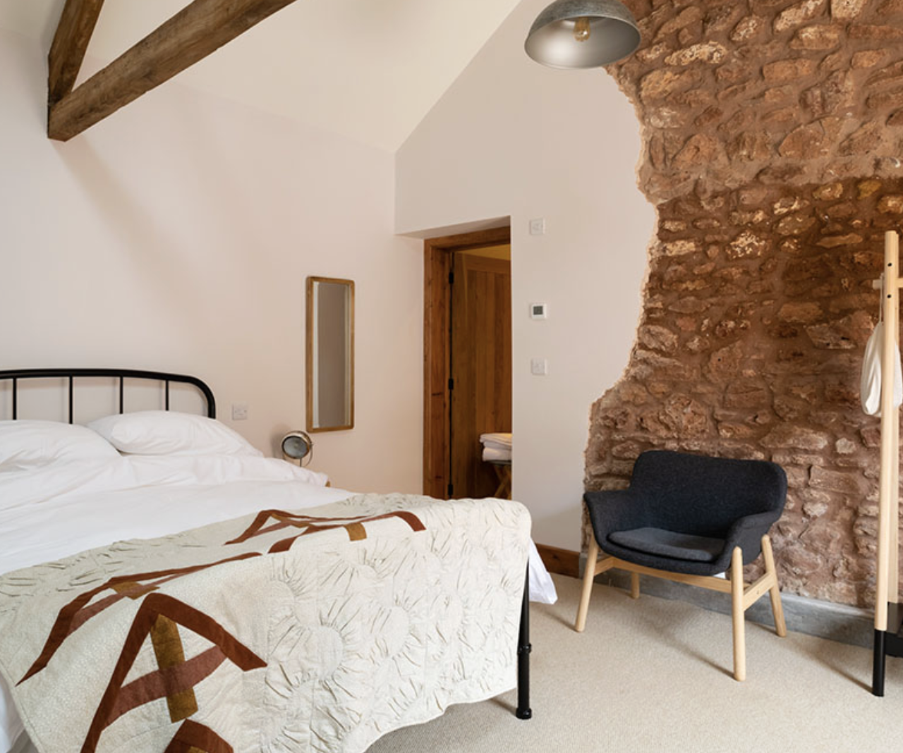 A bedroom with a bed, white linens, and a quilt featuring earthy tones, a black armchair with wooden legs next to a textured red brick wall, and a wooden door leading to a bathroom. There is a small mirror on the wall and a gray ceiling light.