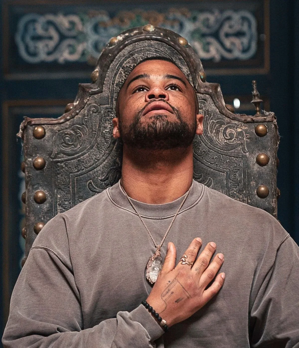 A man with a beard and mustache sitting on a throne-like chair with ornate details, holding his right hand over his chest, looking upwards.