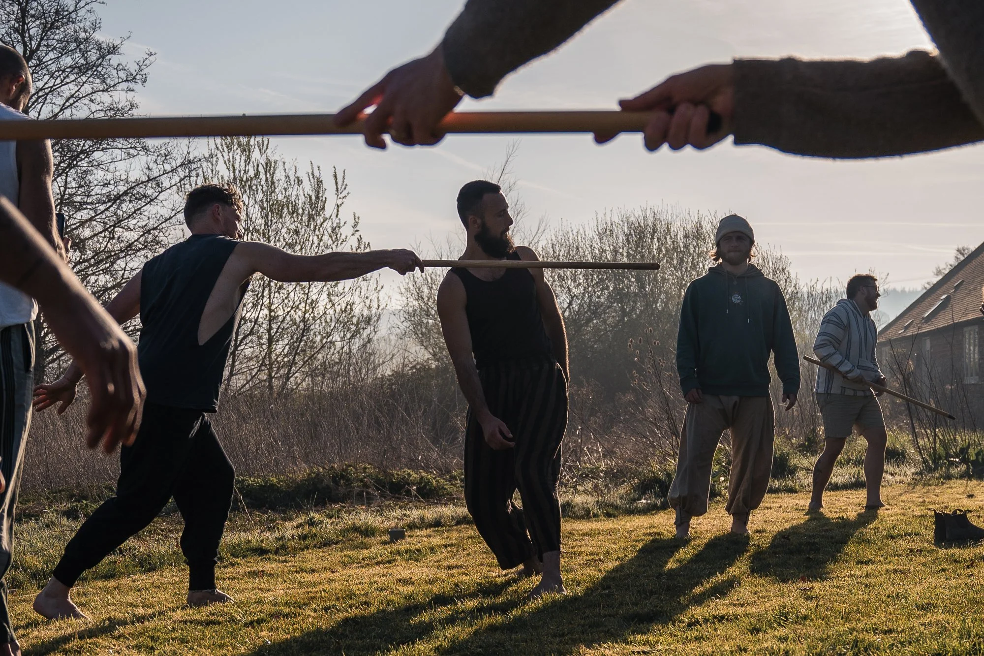 People practicing martial arts outdoors with wooden sticks during sunset, on grassy terrain with trees and a house in the background.