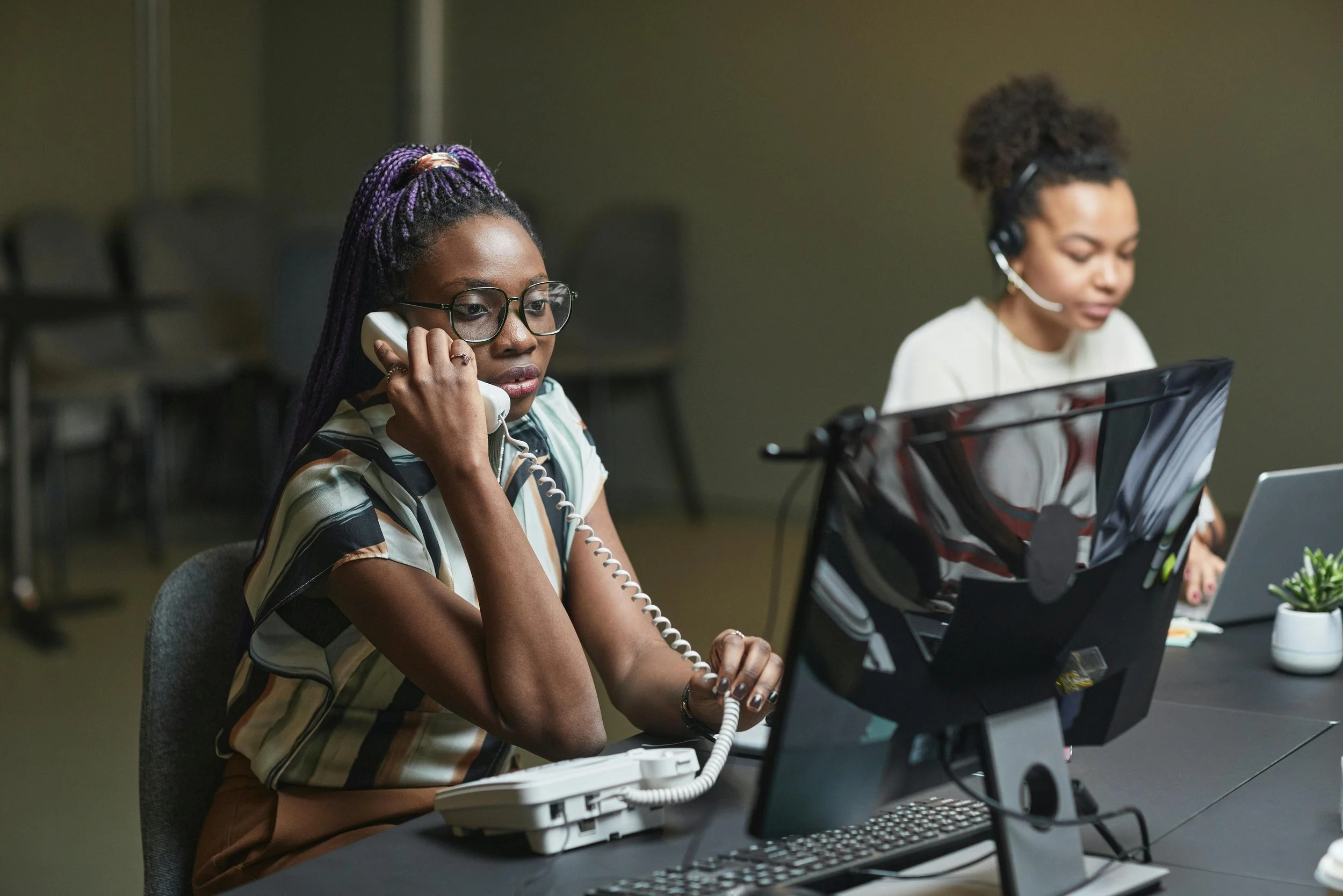 Two women sitting at a desk in an office, both wearing headsets and using computers, one speaking on a landline phone.