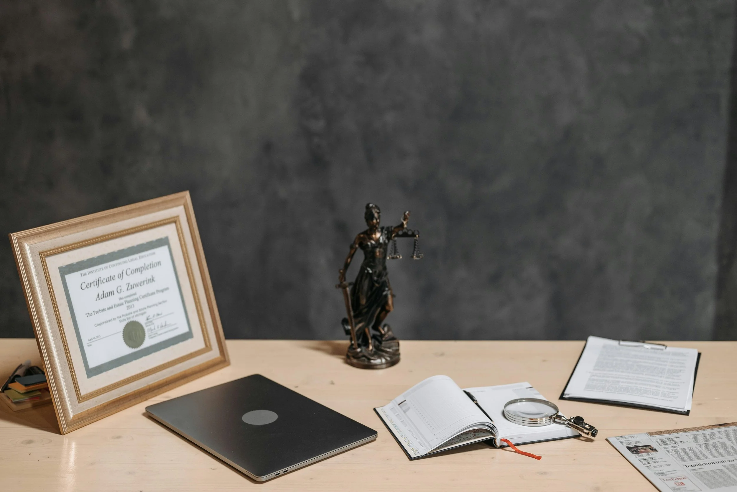 Office desk with a framed certificate, a statue of Lady Justice, a closed laptop, an open planner with a pen, a pair of glasses, and scattered papers against a dark wall background.