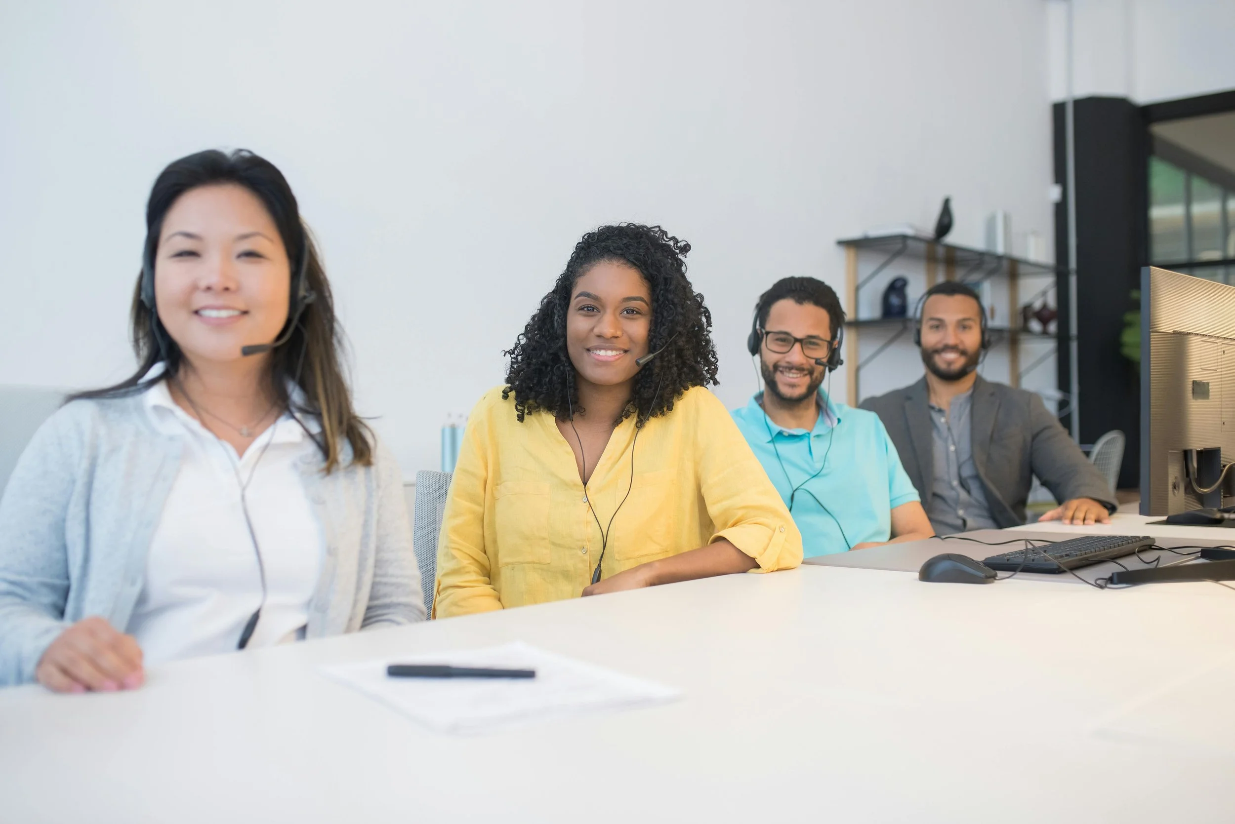 Four diverse individuals sitting at a table in an office, wearing headsets and smiling.