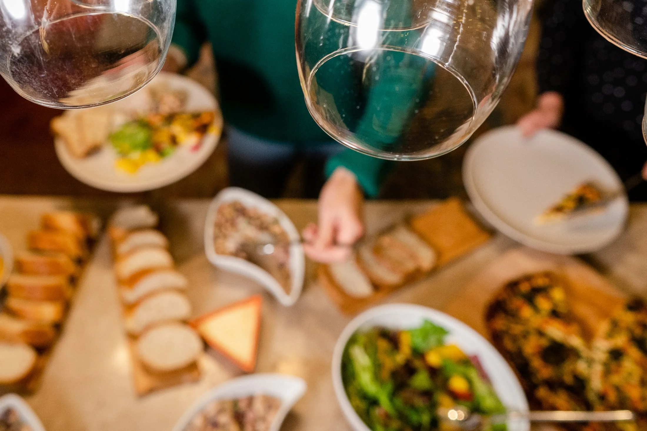 Top-down view of a table with several wine glasses, plates of assorted food including salads, sliced bread, and appetizers, with people reaching for food at a social gathering.