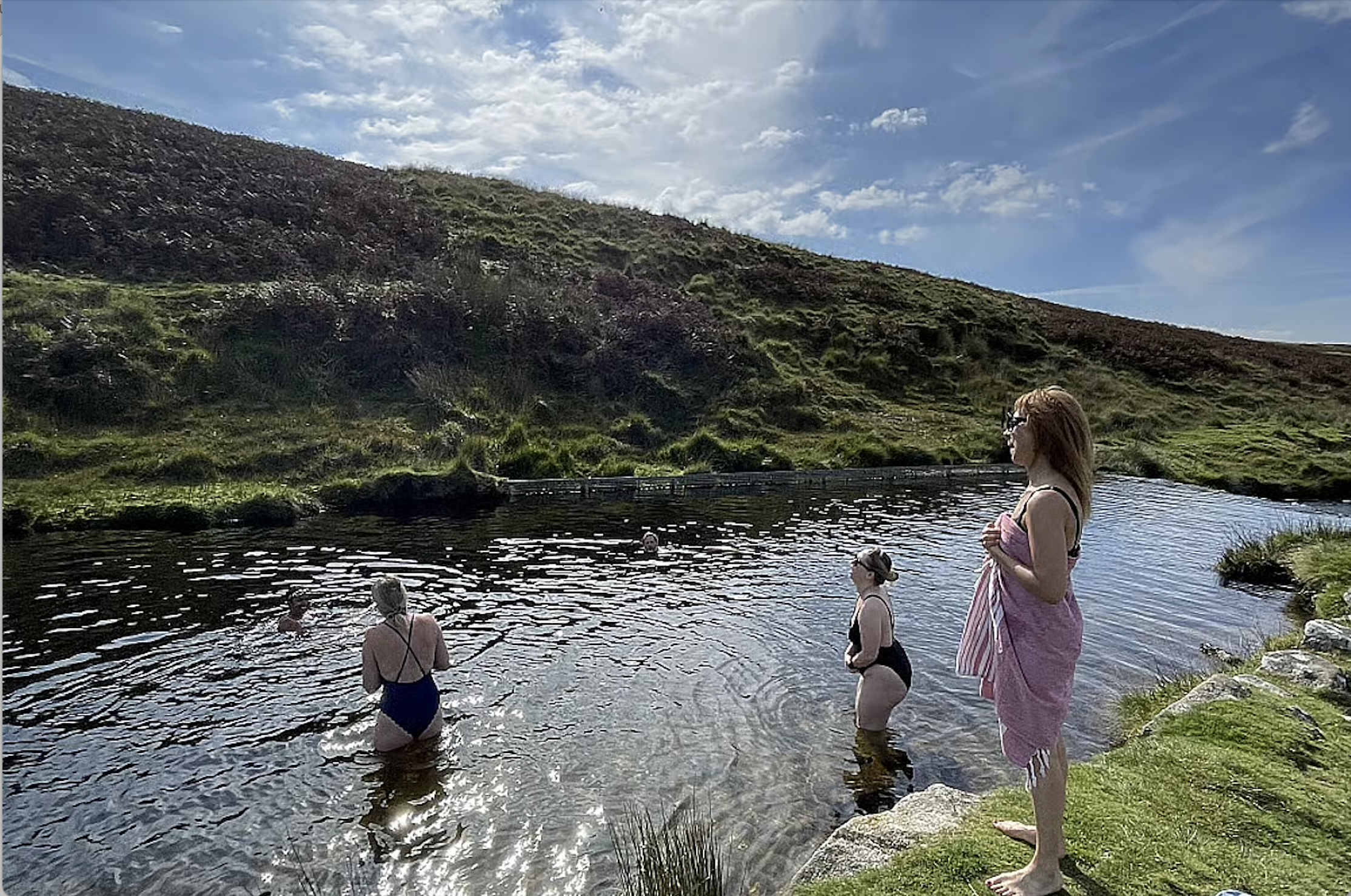 Four women in swimsuits enjoying a small pool in a natural landscape with a grassy hill and a partly cloudy sky.