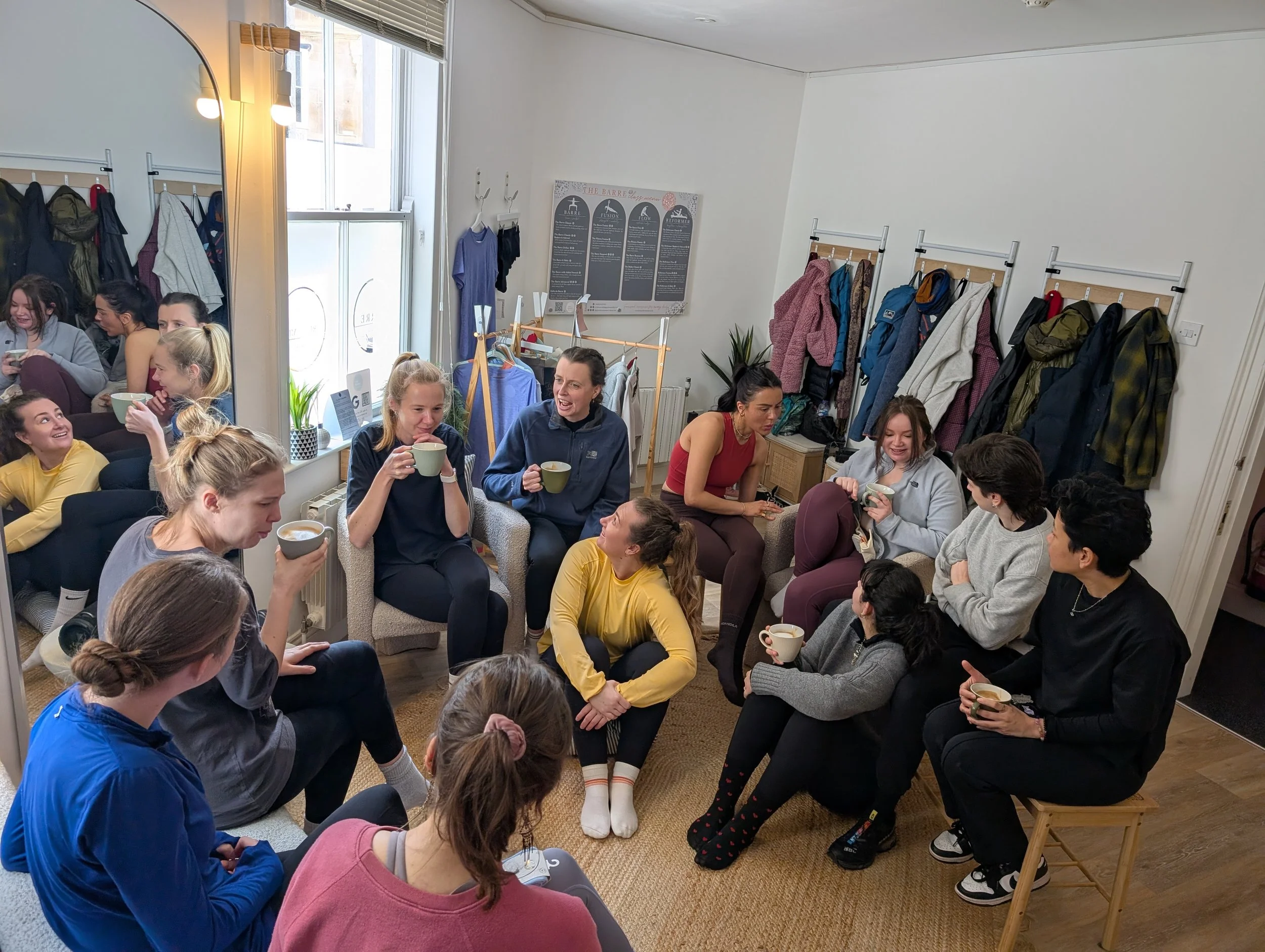 Group of women socializing, sitting on chairs and sofa, in a room with jackets hanging on racks, drinking coffee or tea, enjoying conversation.