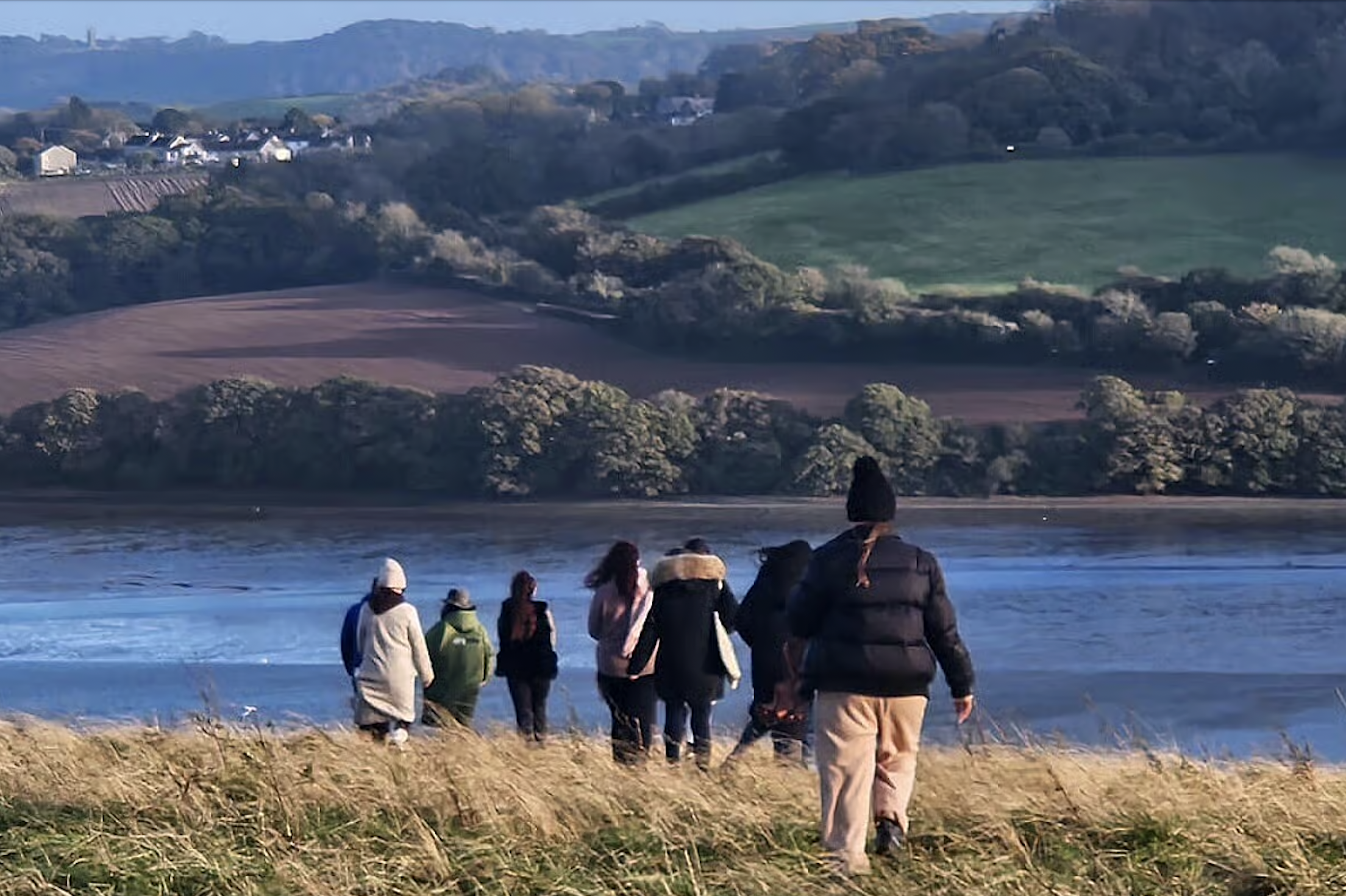 A group of people walking through a grassy field toward a body of water with hills and houses in the background.