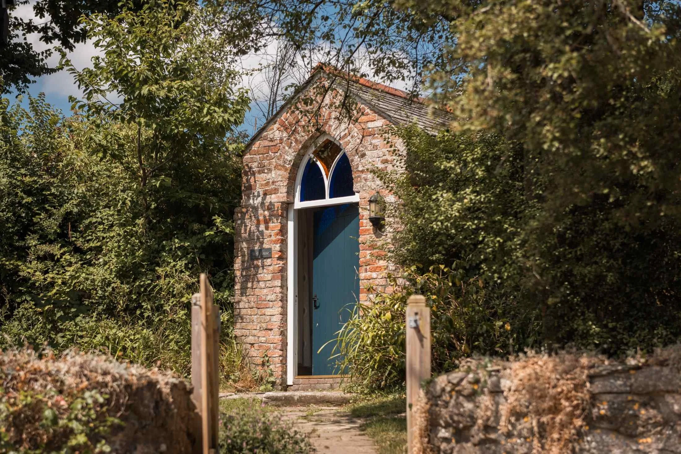 A small brick building with a pointed arch window and blue door, surrounded by lush greenery and trees, with a stone pathway leading to the entrance.