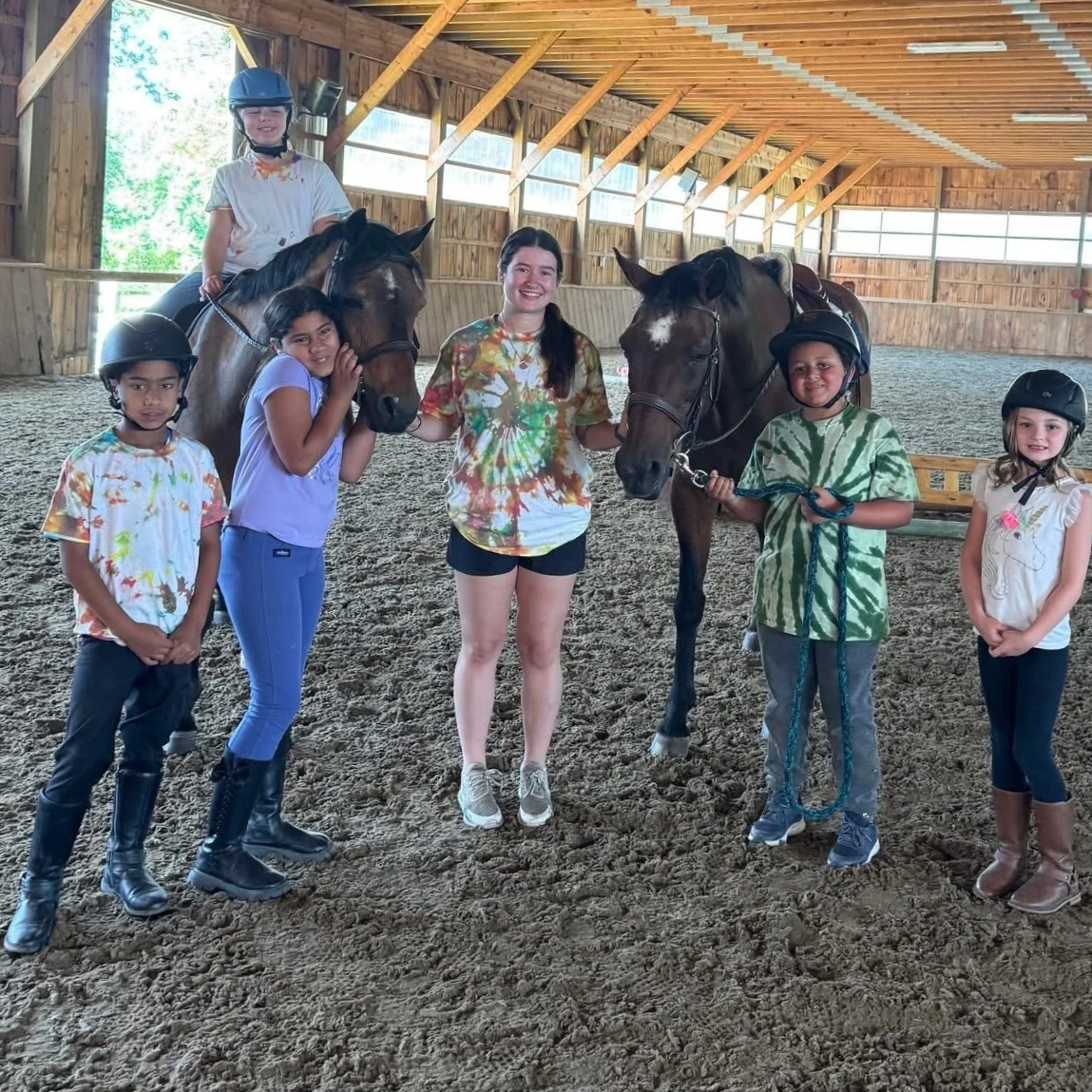 Camp kids smiling with councilor, two brown horses in a horseback riding indoor area.