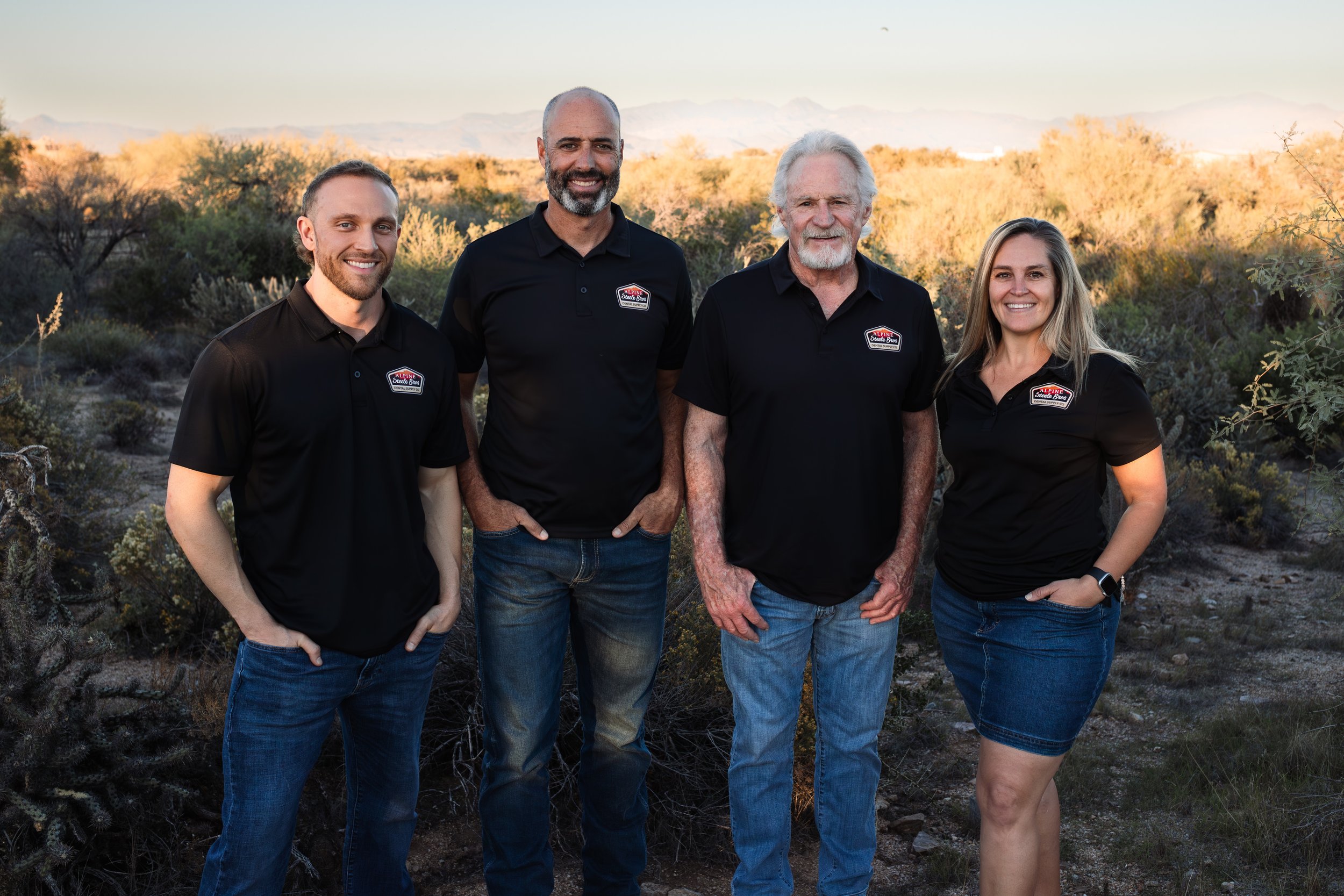 Four people standing outdoors in a desert landscape at sunset, wearing black shirts with a logo, smiling at the camera.