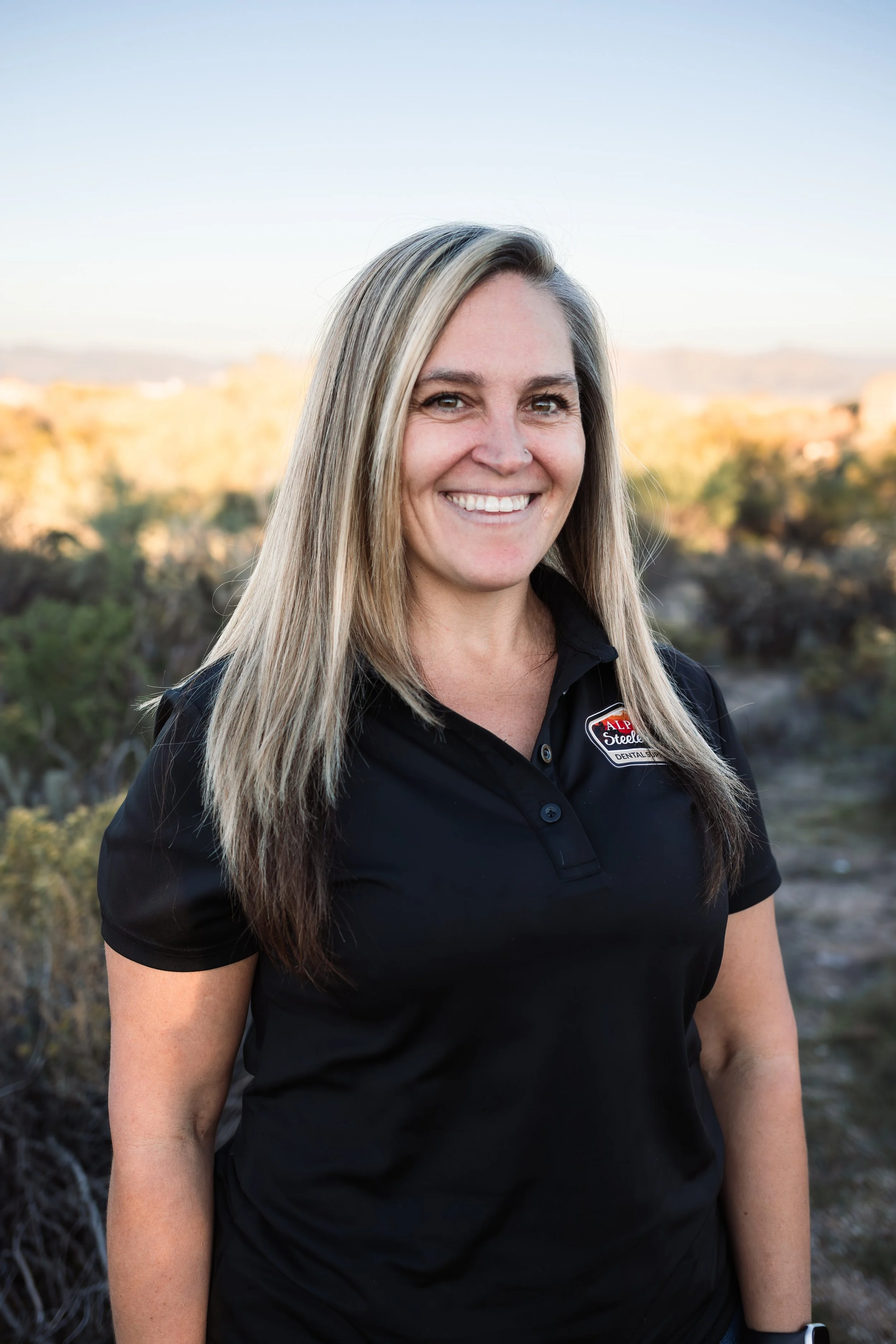 A woman with long blonde hair smiling outdoors, wearing a black polo shirt with a logo that reads 'ALP Steele Dental.' In the background, there are trees and a clear sky.