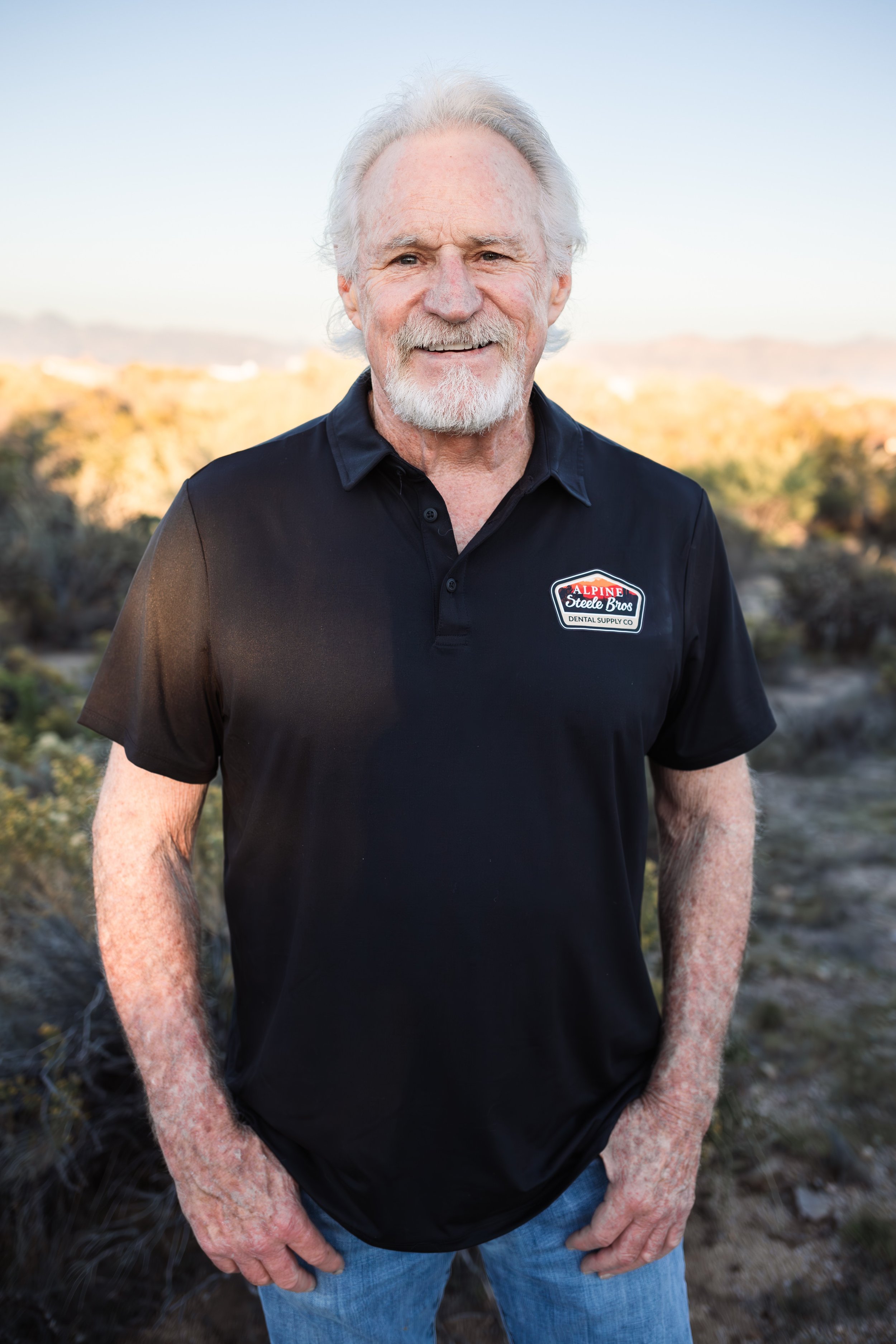 Older man with white hair and beard wearing a black polo shirt with a logo, standing outdoors with a blurred natural landscape background.