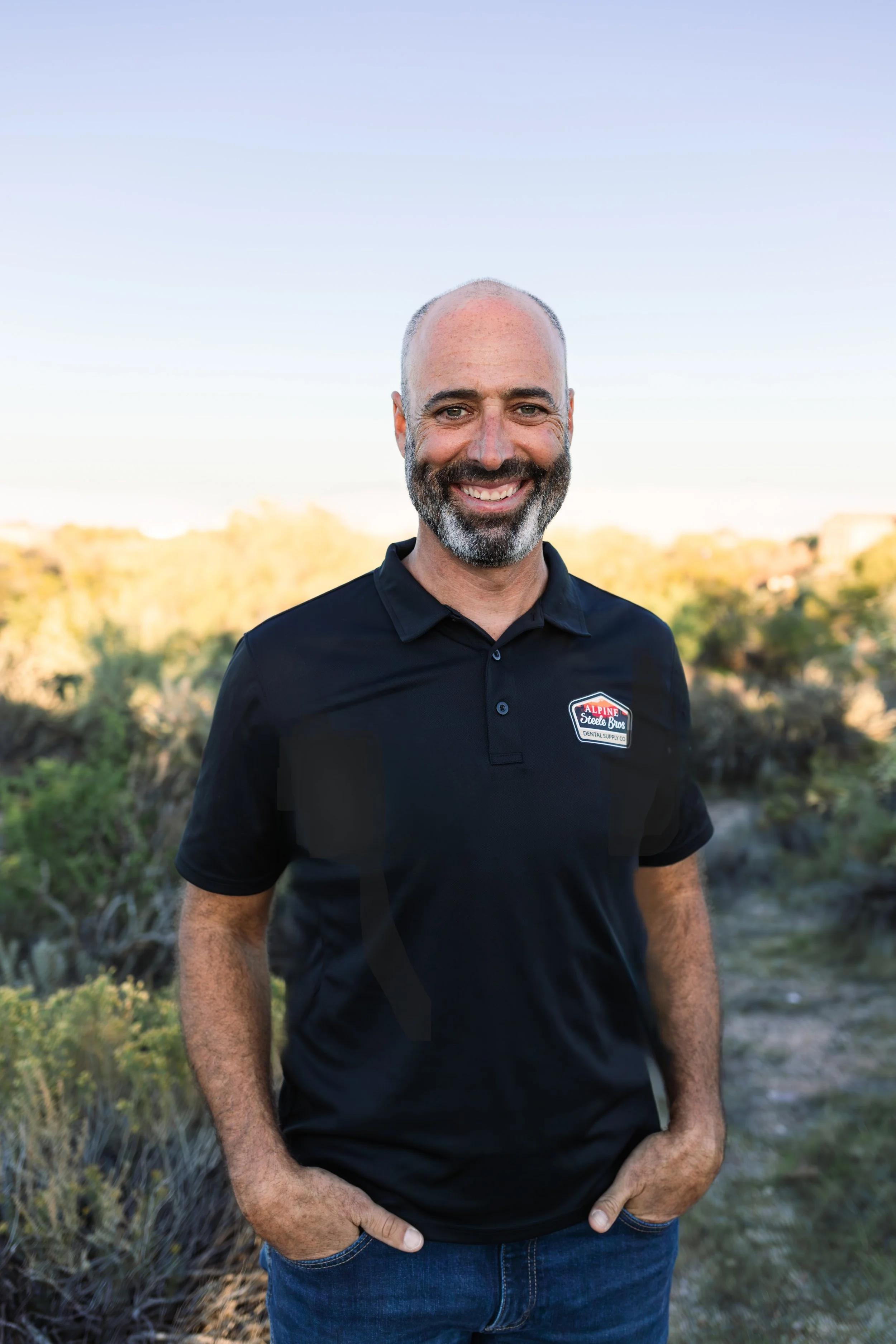 A smiling man in a black polo shirt standing outdoors with a natural landscape in the background.