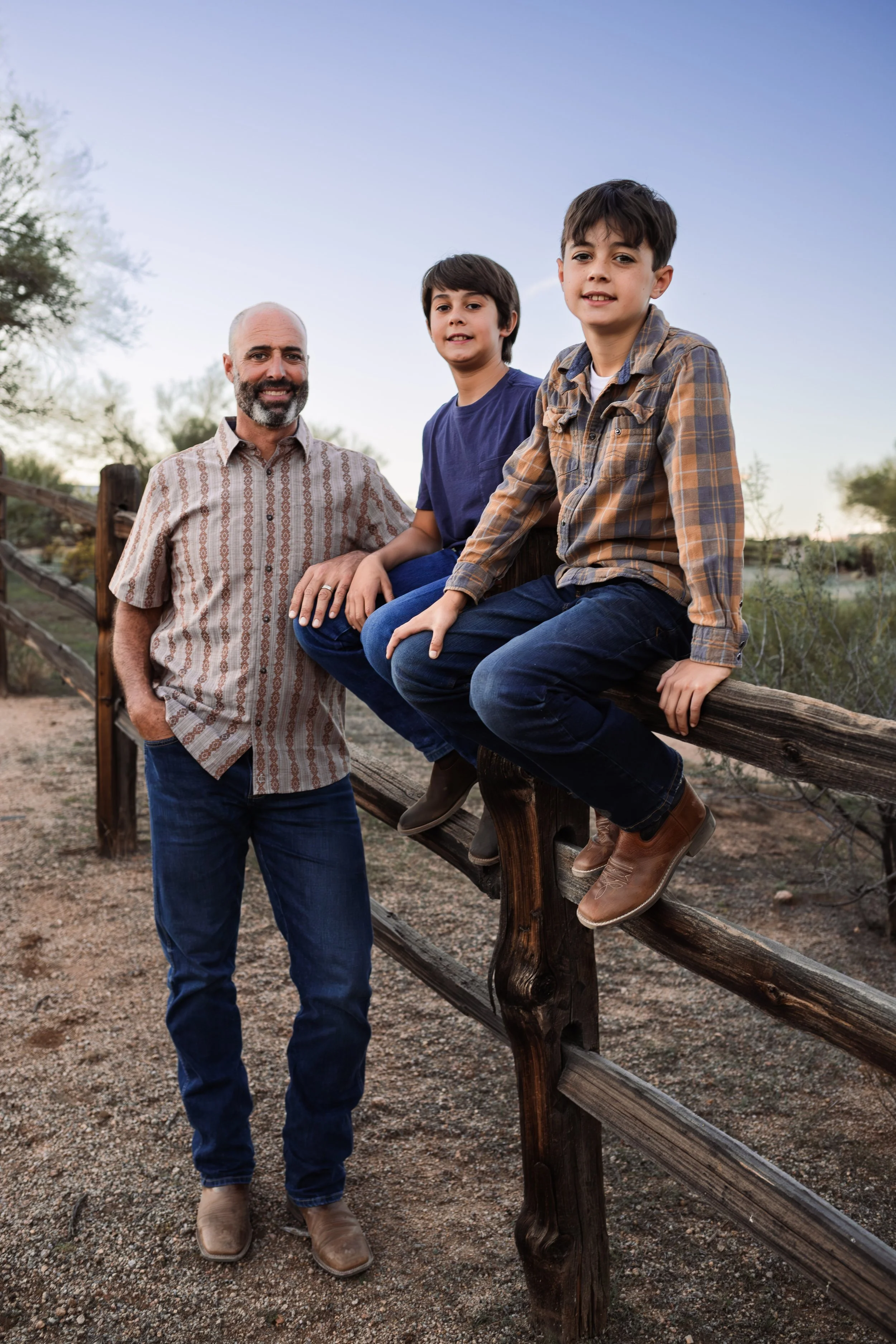 A father with two young sons outdoors on a wooden fence at sunset with trees in the background.