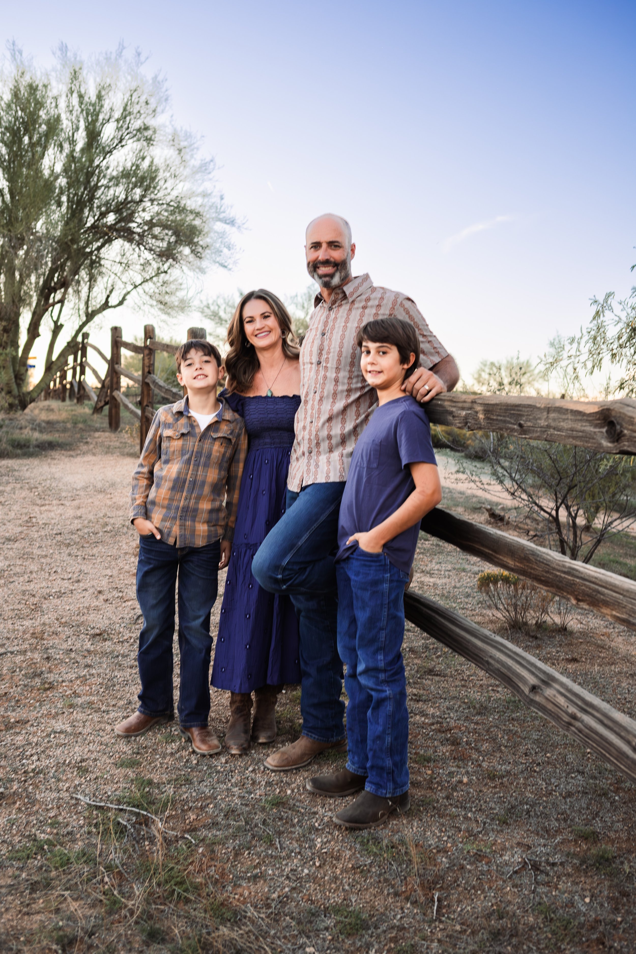 Family of four outdoors in a desert-like setting, smiling, with a wooden fence and trees in the background, during sunset.