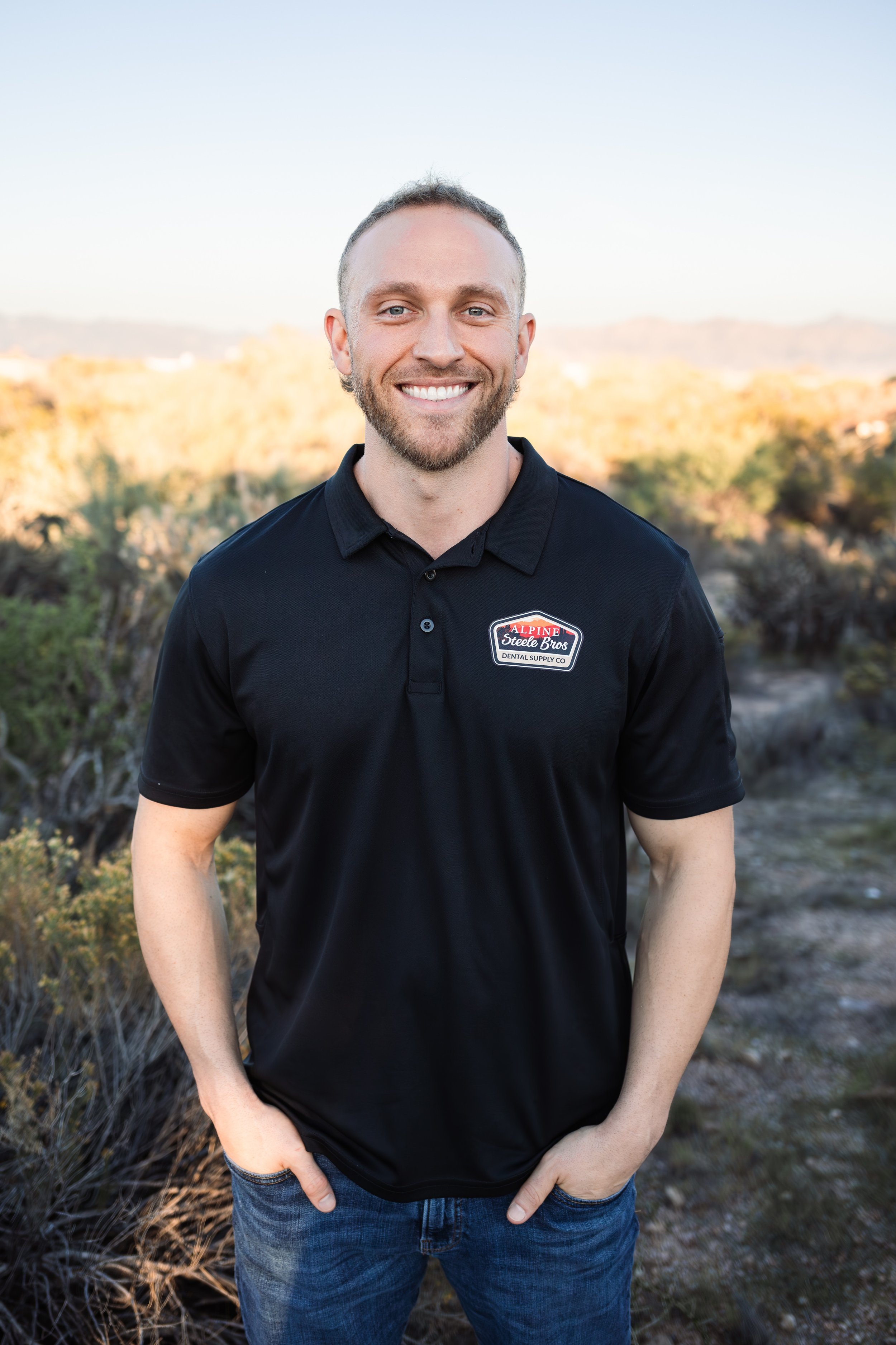 Smiling man outdoors in a natural setting, wearing a black polo shirt with a company logo, hands in pockets, with a backdrop of trees and hills under a clear sky.