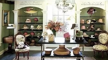 A vintage-style room with two green bookshelves filled with hats and accessories, a mirror on the left, a window in the center with a chandelier overhead, and a table in front holding various hats and decorative items, flanked by antique chairs.
