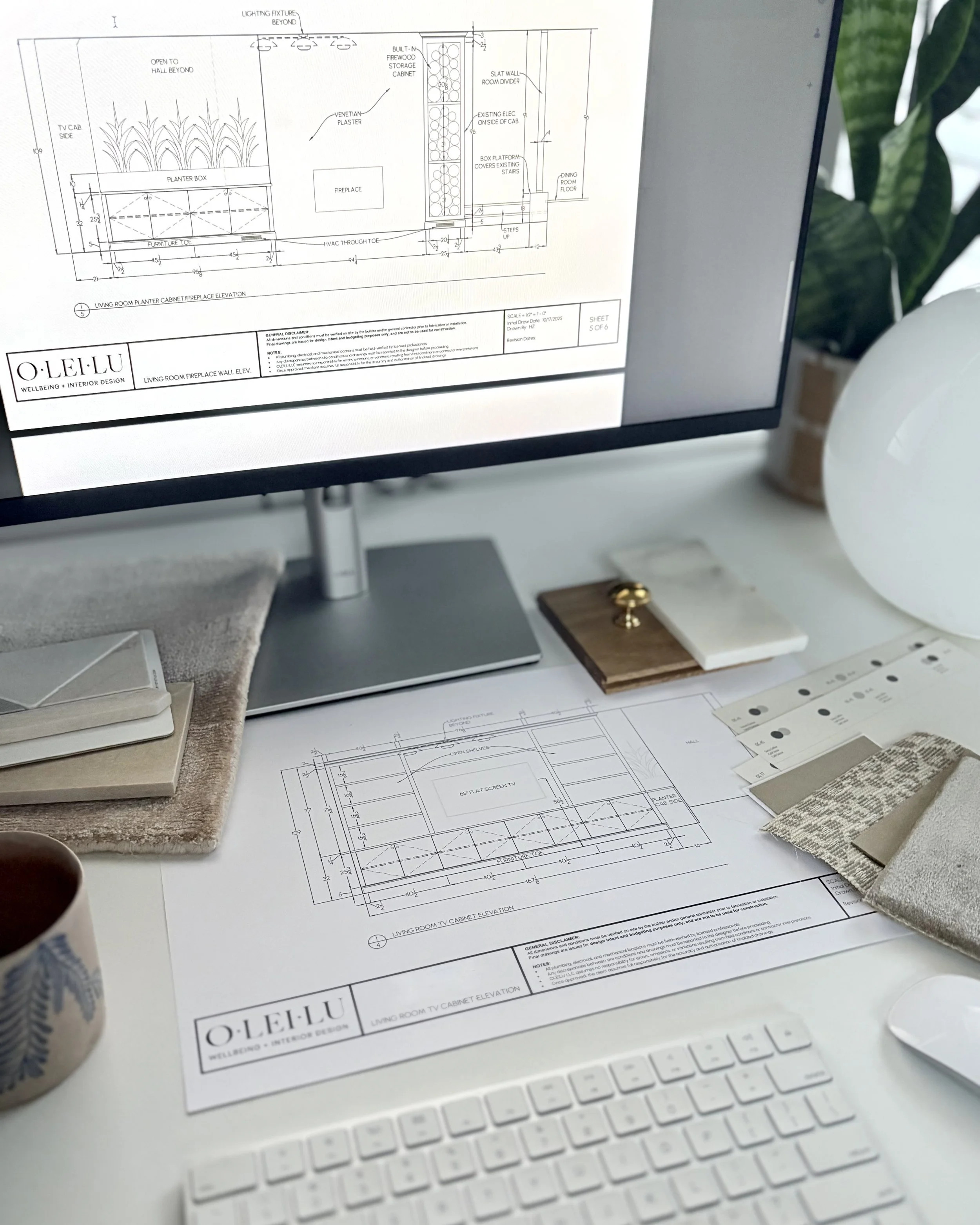 Architectural blueprints and interior design sketches on a workspace desk with a computer monitor, fabric swatches, color sample cards, a mug, and decorative objects, with a plant in the background.
