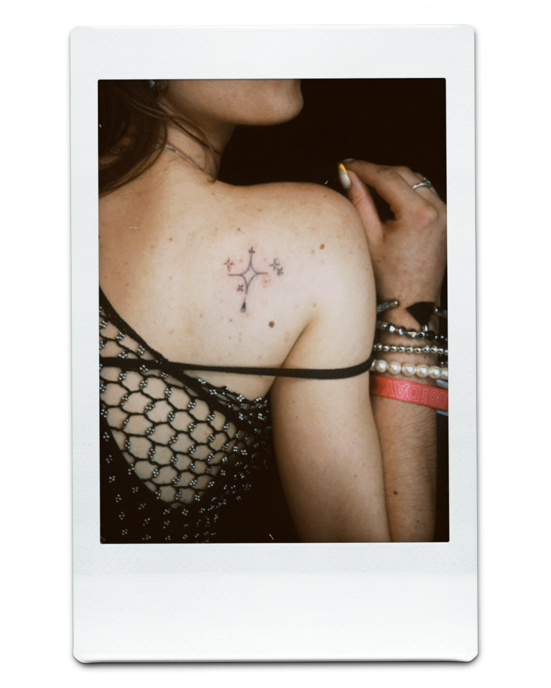 Person wearing a netted top, showing a shoulder tattoo of a stylized cross with floral details, accessorized with bracelets.