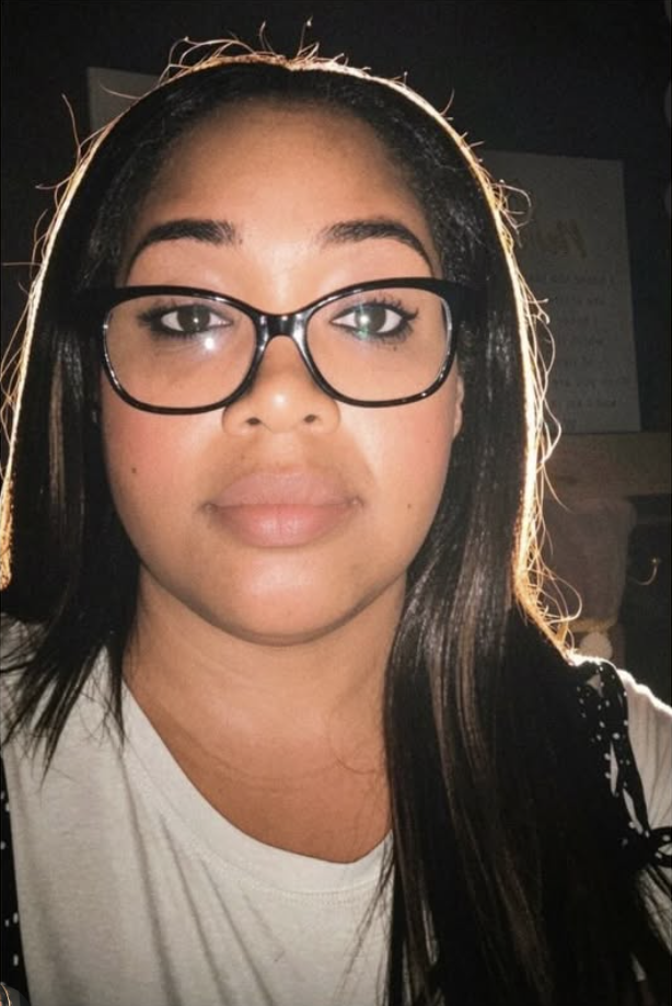A young woman with long dark hair, wearing glasses with black frames, and a white top. She has a neutral expression and is looking directly at the camera, with a dark background and some light highlighting her hair.