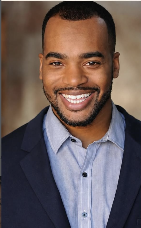 Headshot of a smiling man with short black hair, a beard, and brown skin, wearing a light blue collared shirt and a dark blazer.