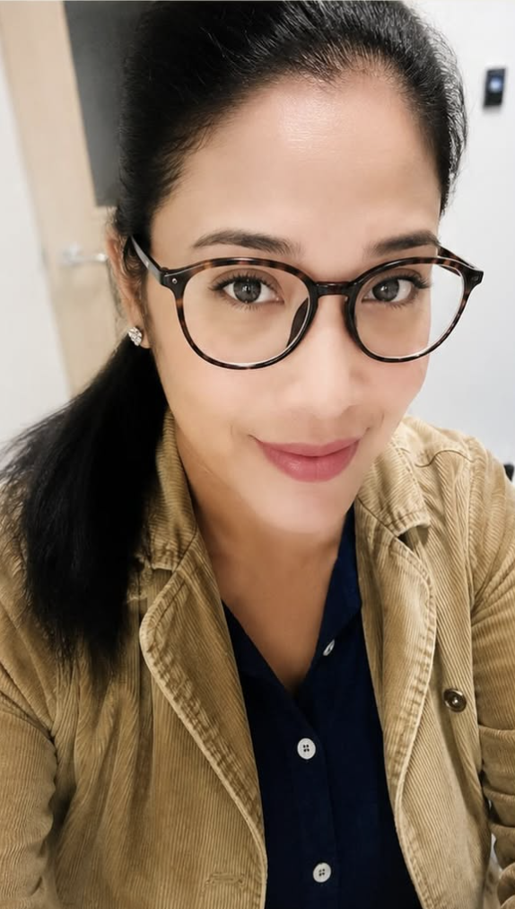 Close-up of a smiling woman with black hair, wearing tortoiseshell glasses, earrings, a navy blue shirt, and a tan jacket indoors.