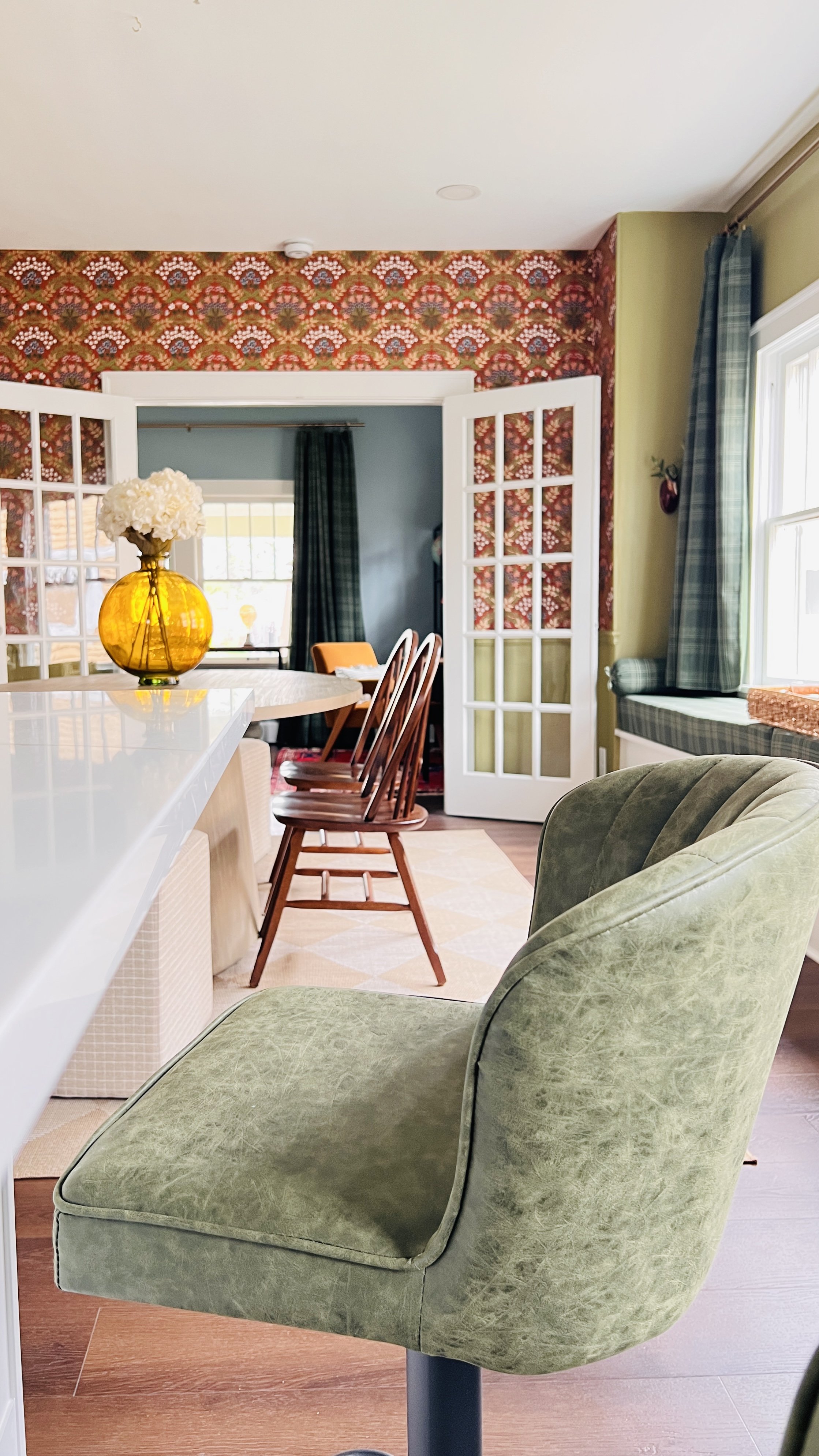 Interior of a room with a green upholstered bar stool in the foreground, a yellow vase with white flowers on a countertop, French doors leading to another area, colorful patterned wallpaper, and wooden chairs around a round table.