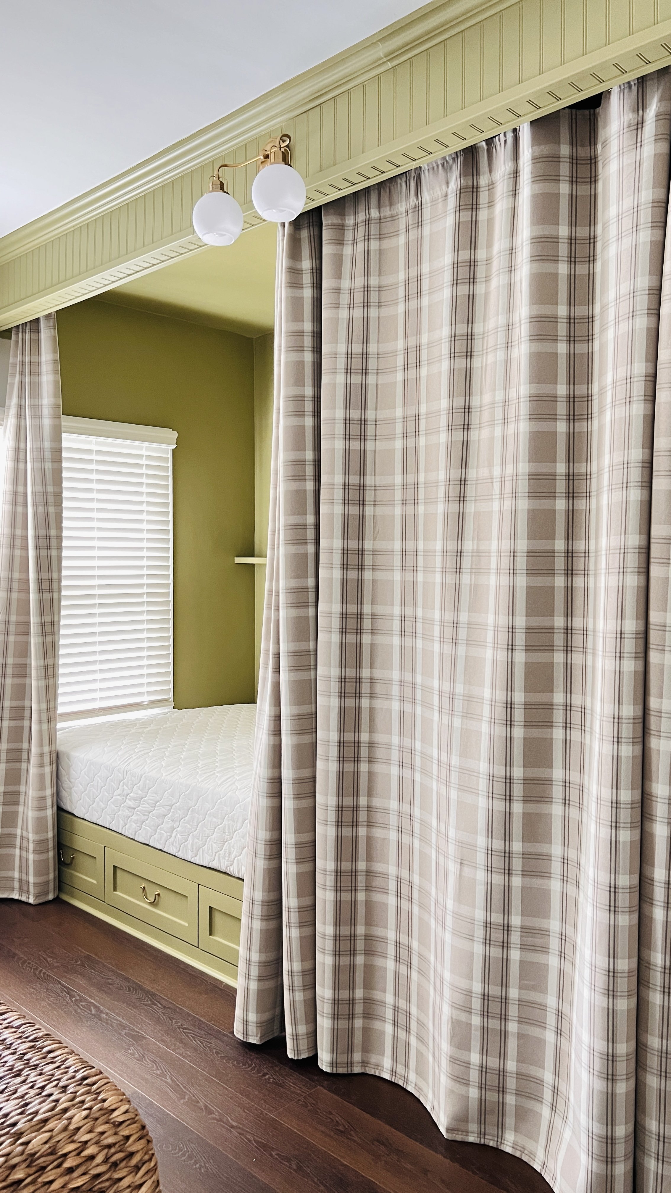 A cozy napping nook with a daybed, plaid curtains, and green walls, featuring white blinds and a wooden floor.