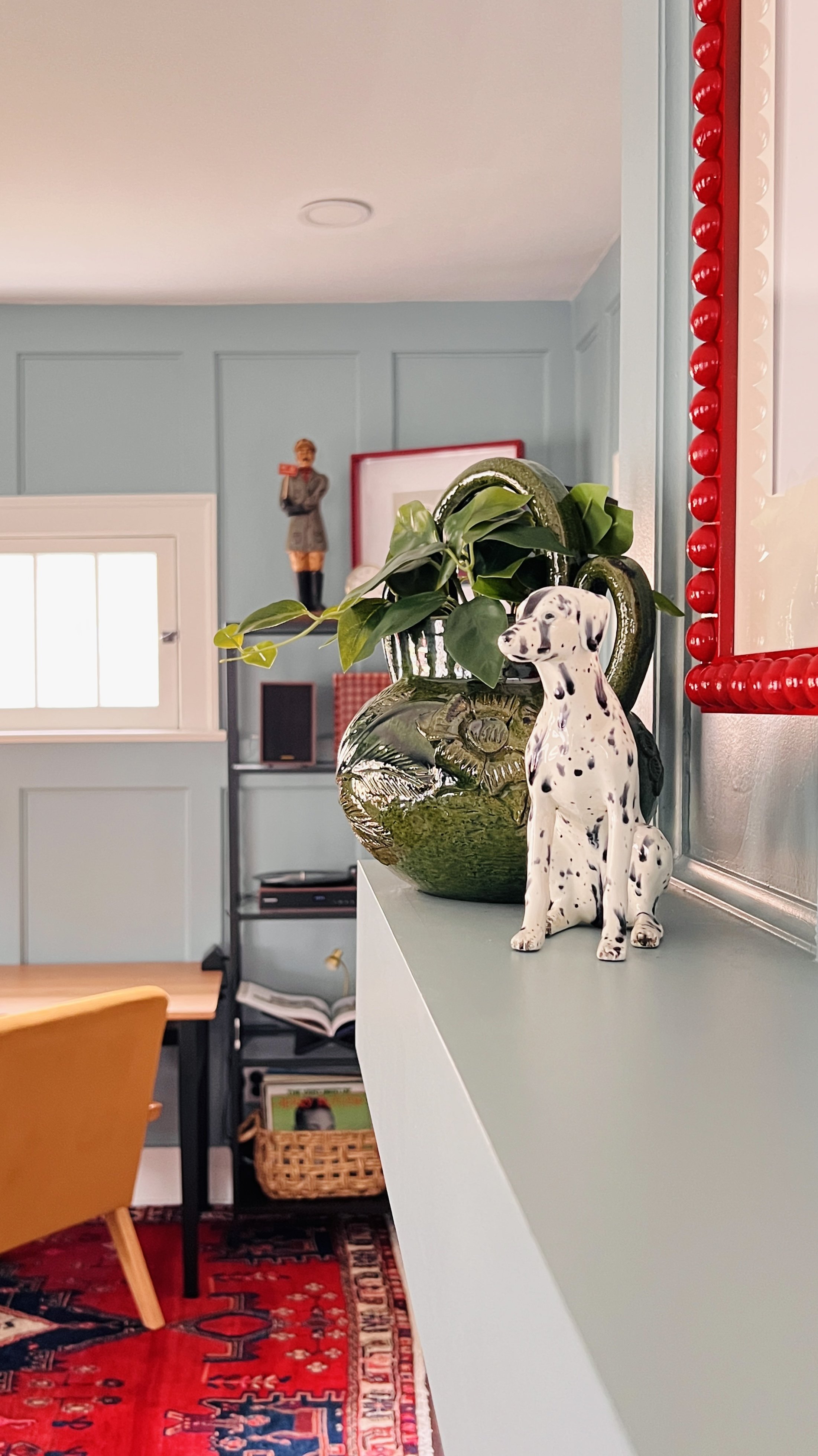 Interior room with green walls, featuring a ceramic Dalmatian dog statue on a shelf next to a green plant in a vase, a chair with a red patterned rug, and a small window.