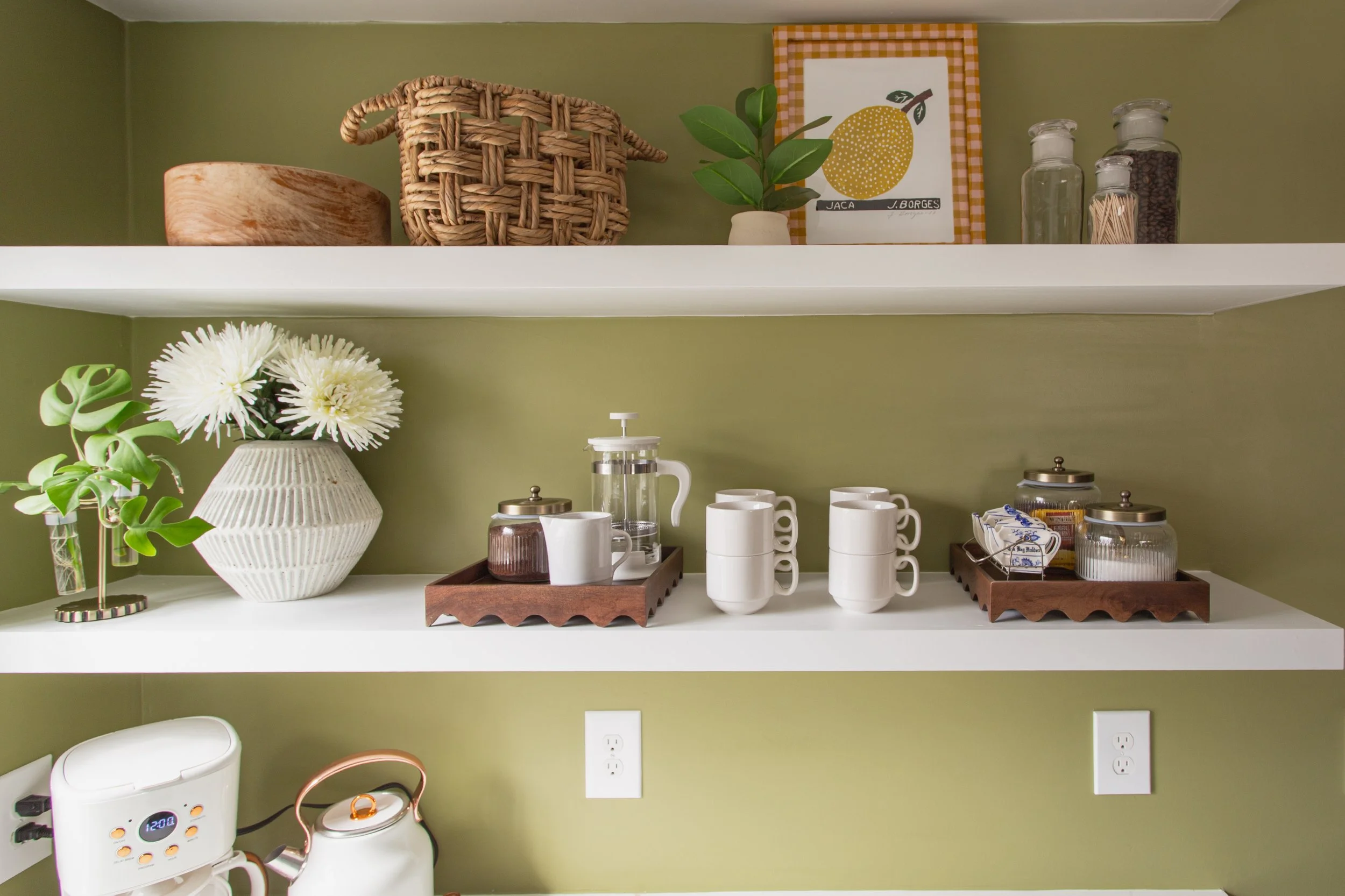 Kitchen shelves with green wall background, featuring a woven basket, a plant, a framed lemon print, vases with white flowers, a French press, mugs, and glass jars with coffee beans and sugar.