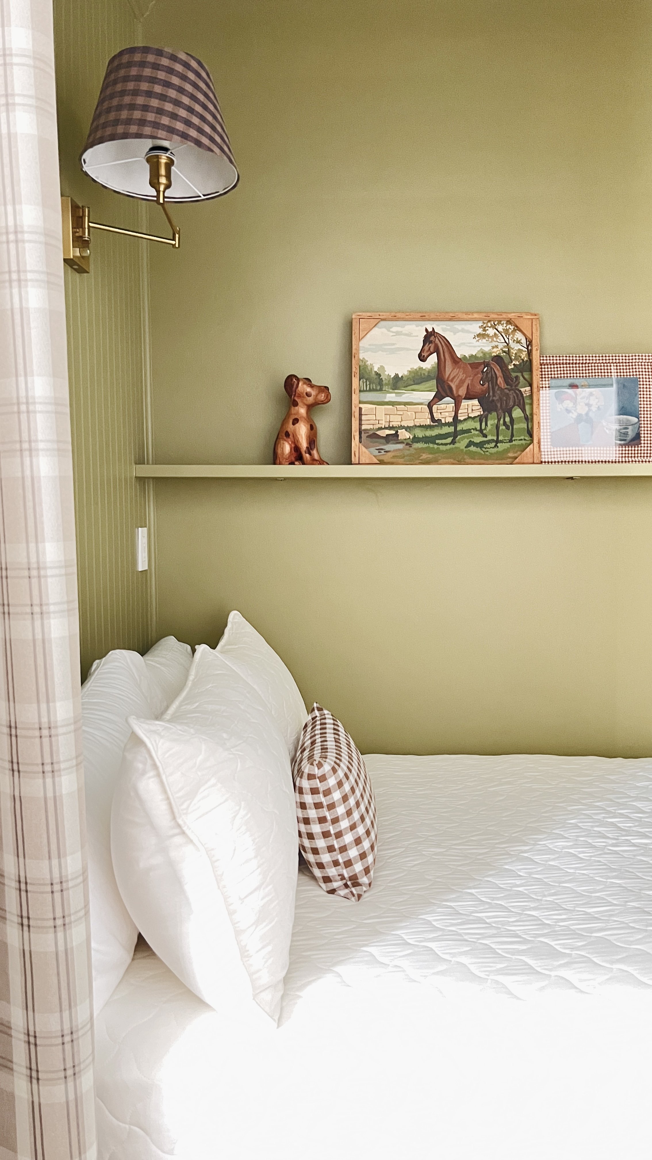 Cozy bedroom with green walls, a wall sconce with a plaid lampshade, a shelf displaying a horse painting and dog figurine, a white bed with white pillows, and a plaid accent pillow.