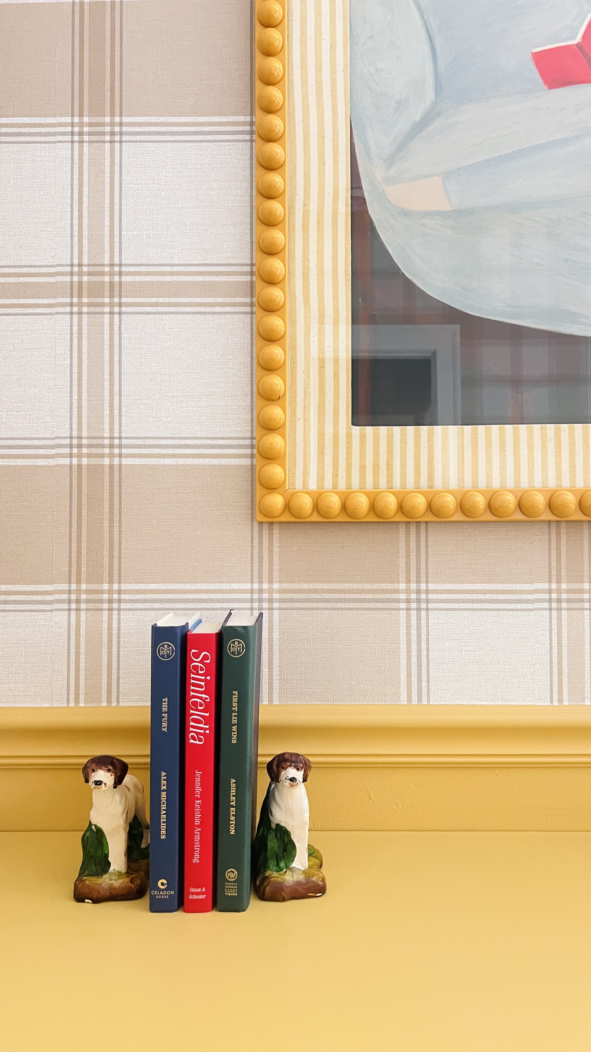 Books on a yellow shelf with dog-shaped bookends, a plaid wallpaper background, and a framed painting with a yellow beaded frame.