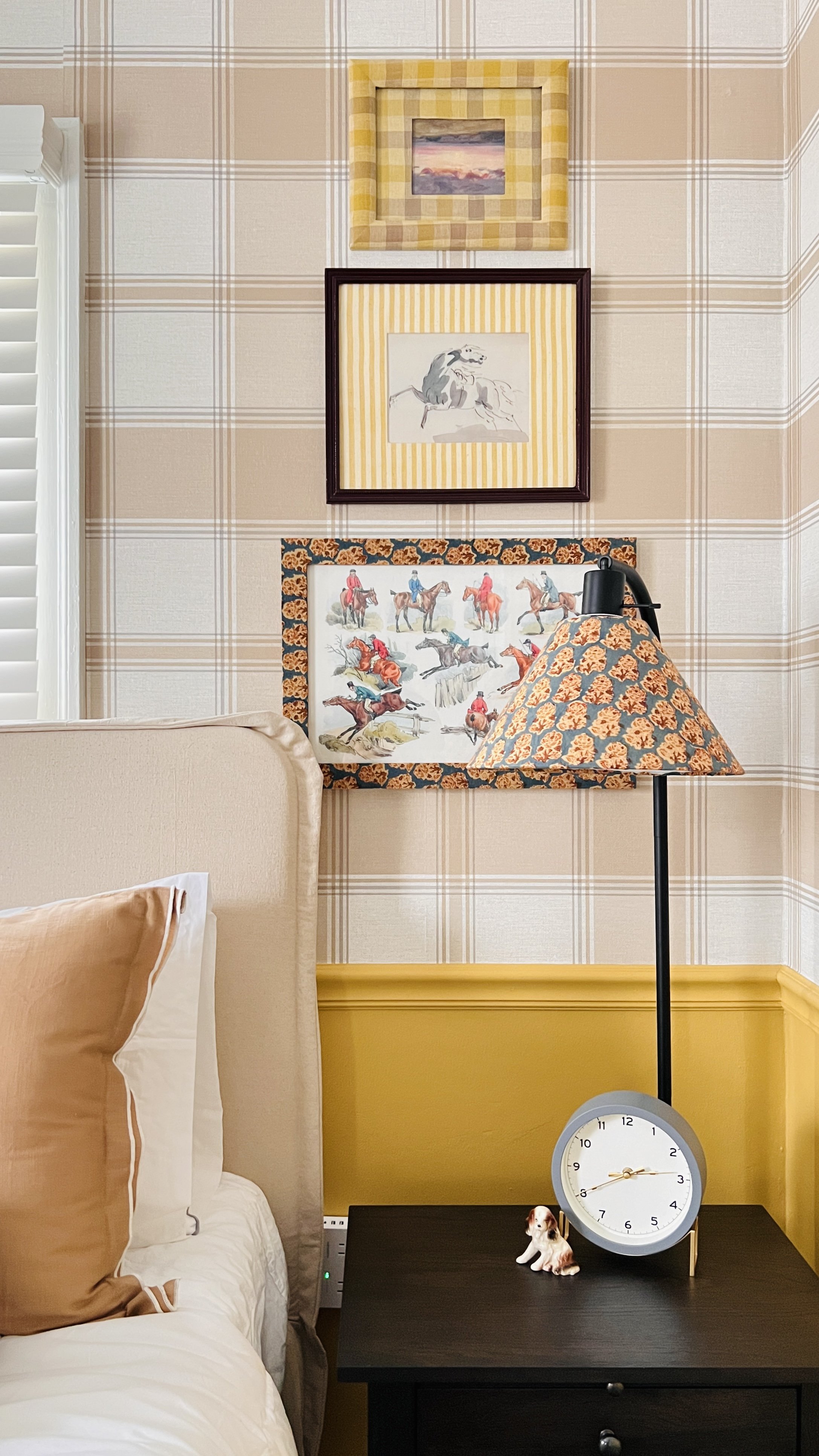 Bedroom scene with beige plaid wallpaper, framed artwork, and a beige bed with pillows. A black nightstand holds a round clock and a small dog figurine, and a lamp with a patterned shade sits on the table.