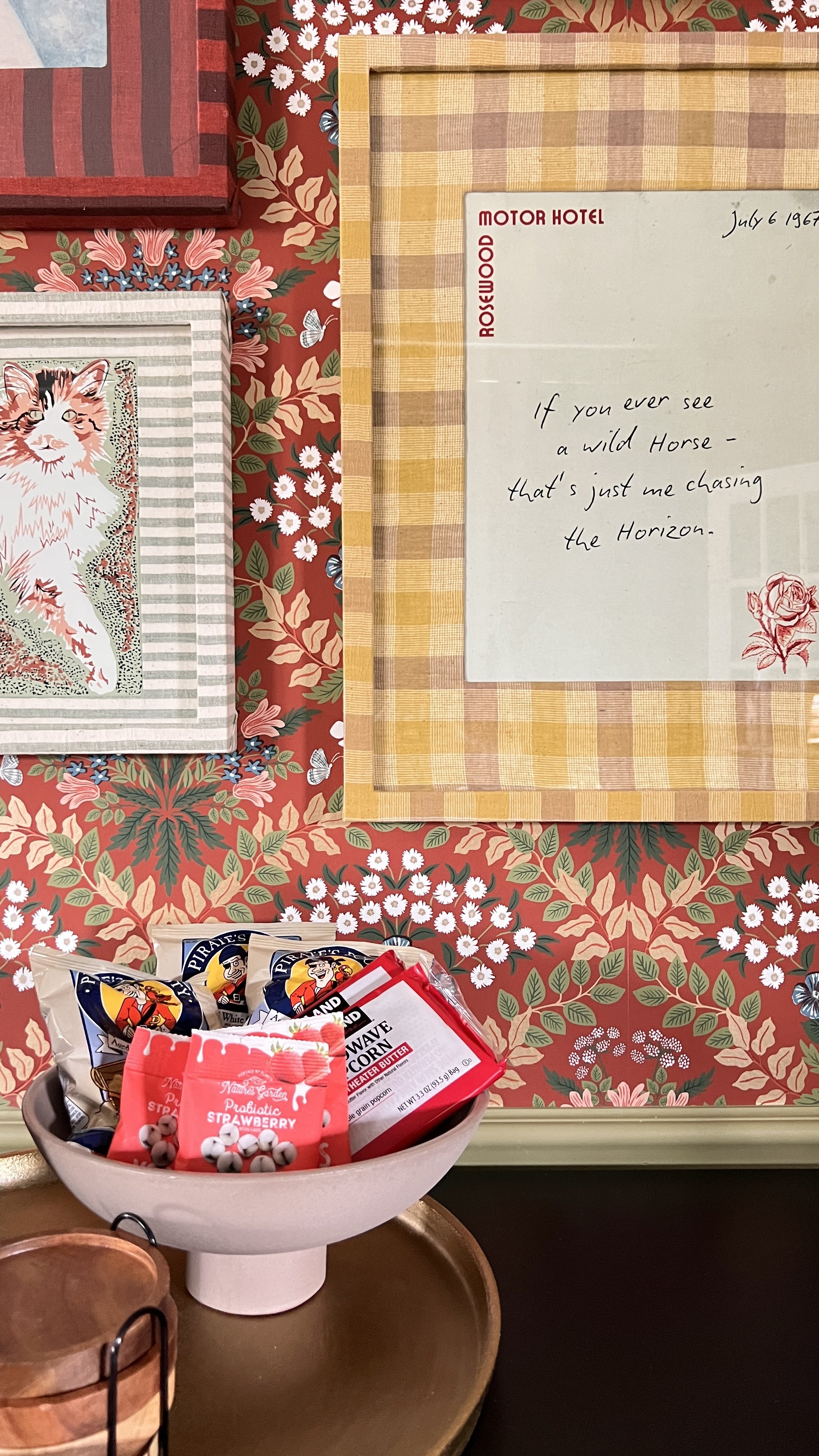 A decorated wall with floral wallpaper and framed artworks, including a handwritten note. In front, a bowl holds snack packets such as Pirate's Booty, microwavable popcorn, and dried strawberries.