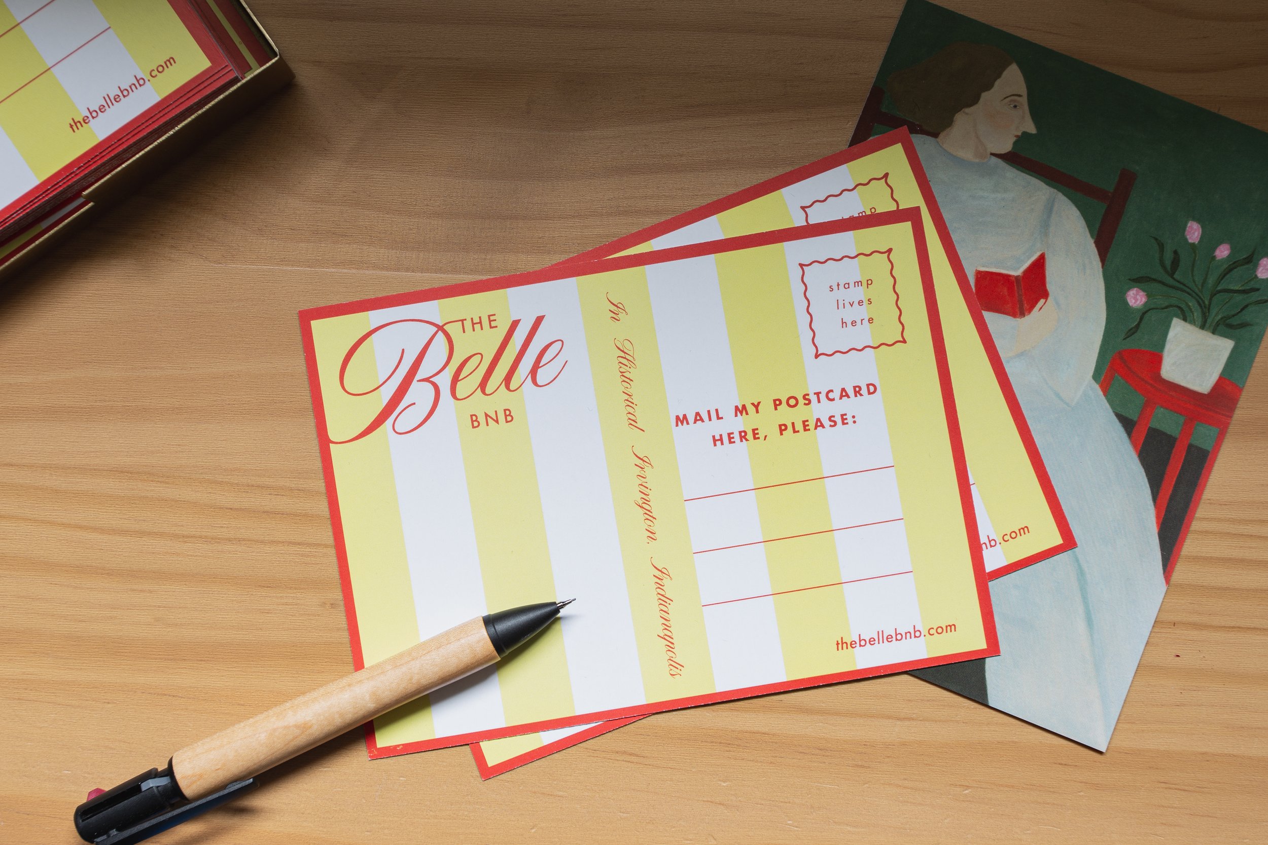 Postcards from The Belle BNB on a wooden table with a pen. The postcards feature yellow and white stripes, contact info, and space for a stamp. A decorative card with an illustration of a person reading on a chair is also visible.
