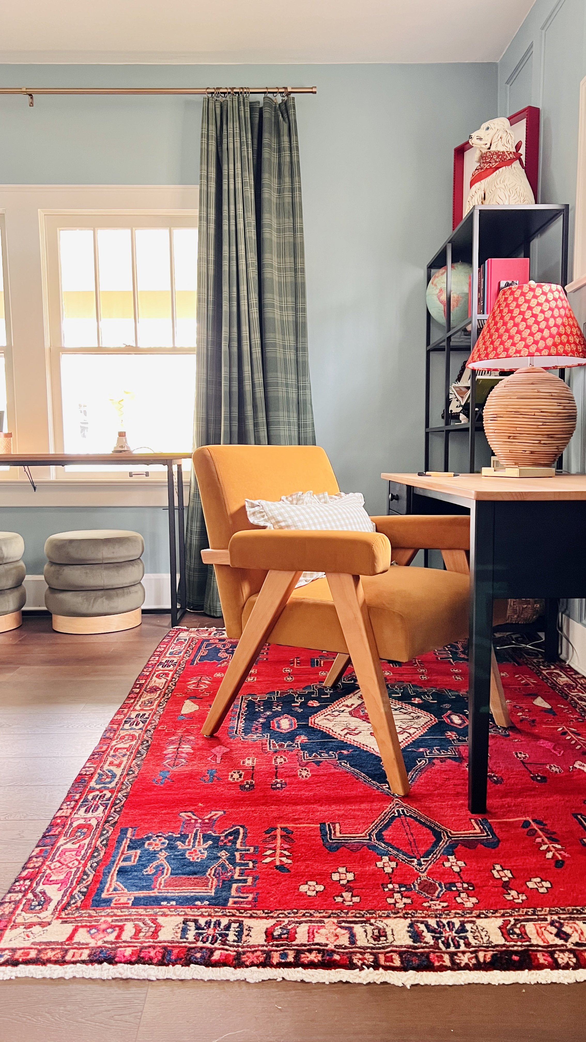 A cozy home office with a yellow armchair and decorative pillows, set on a vibrant red patterned rug. A wooden desk with a red and beige lamp is present, alongside a black metal shelving unit with books and a globe. The room features plaid curtains a