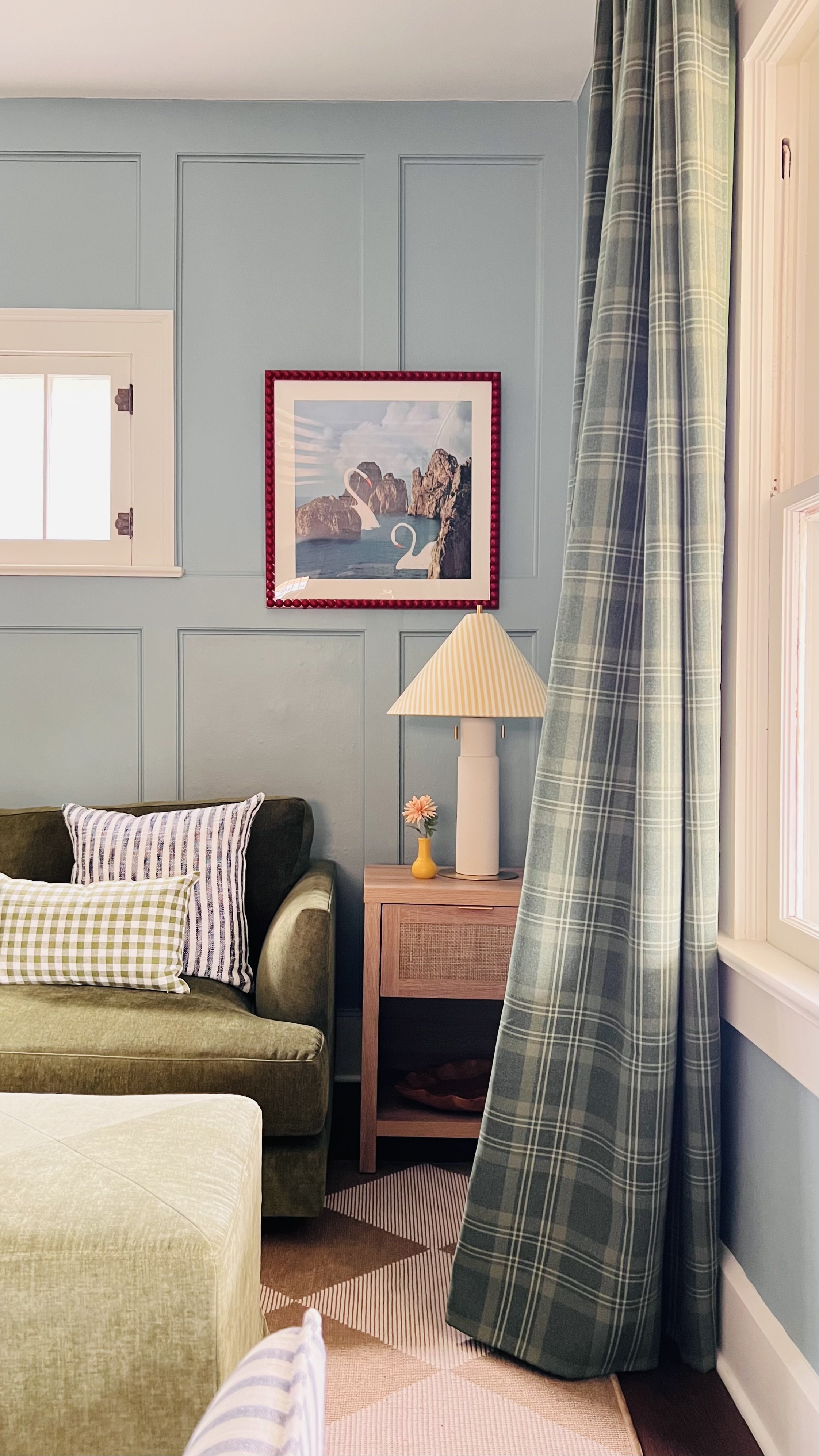 Cozy living room corner with a green velvet sofa, plaid curtains, framed wall art, and a wooden side table with a striped lamp and a small flower in a vase.