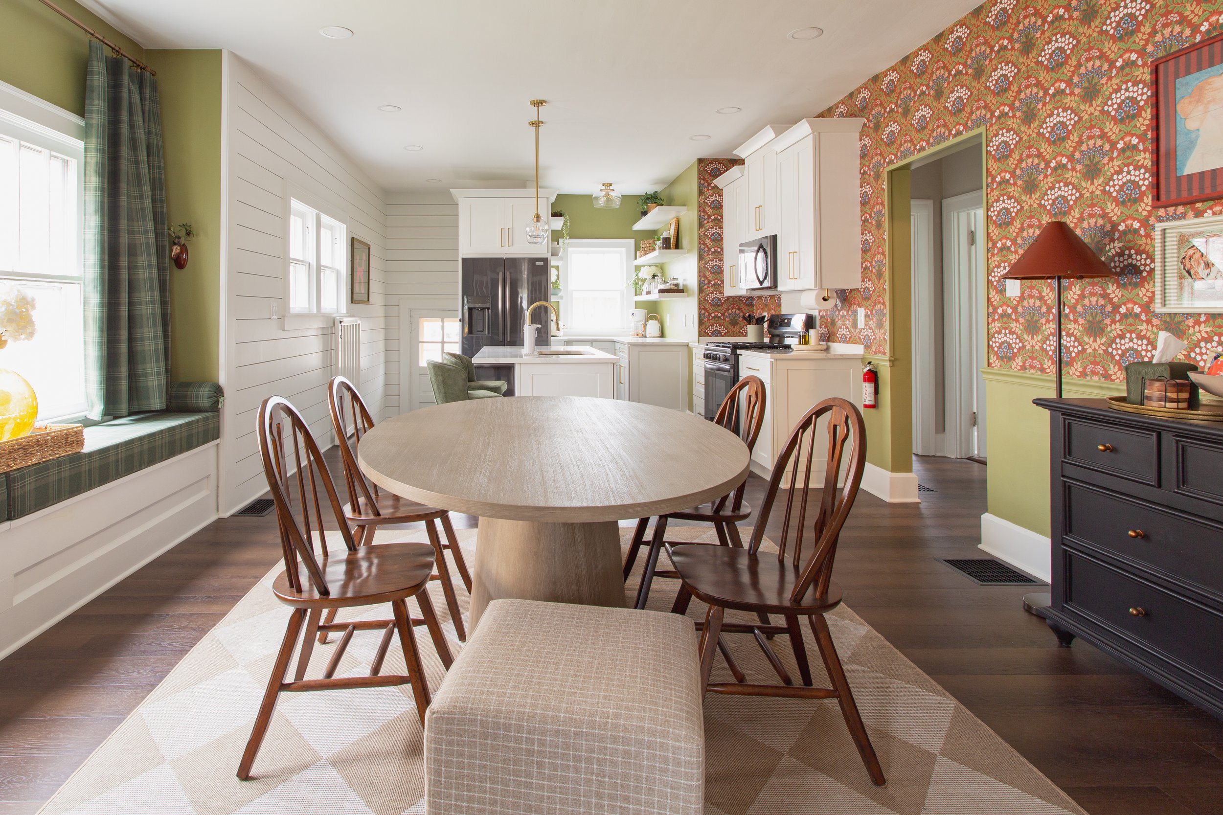 A cozy kitchen and dining area with white cabinets, an orange and green floral patterned accent wall, a wooden dining table with matching chairs, a window seat with green plaid cushions, hardwood floors, and modern appliances.
