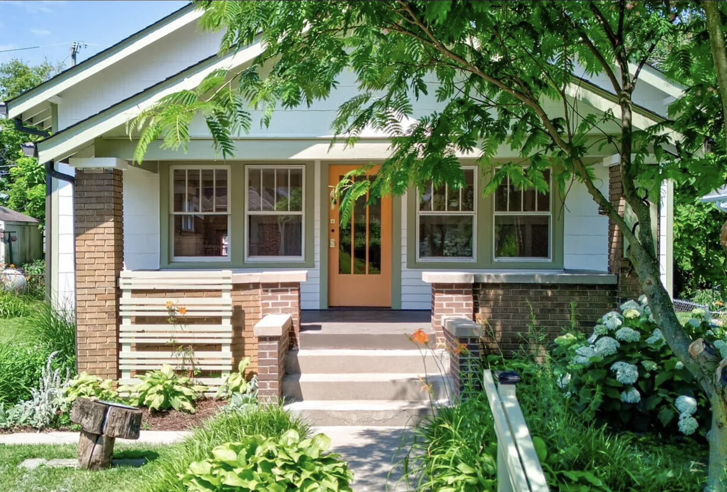 Charming craftsman-style bungalow with a porch, surrounded by lush greenery and flowers, featuring a brick base and light-colored siding.