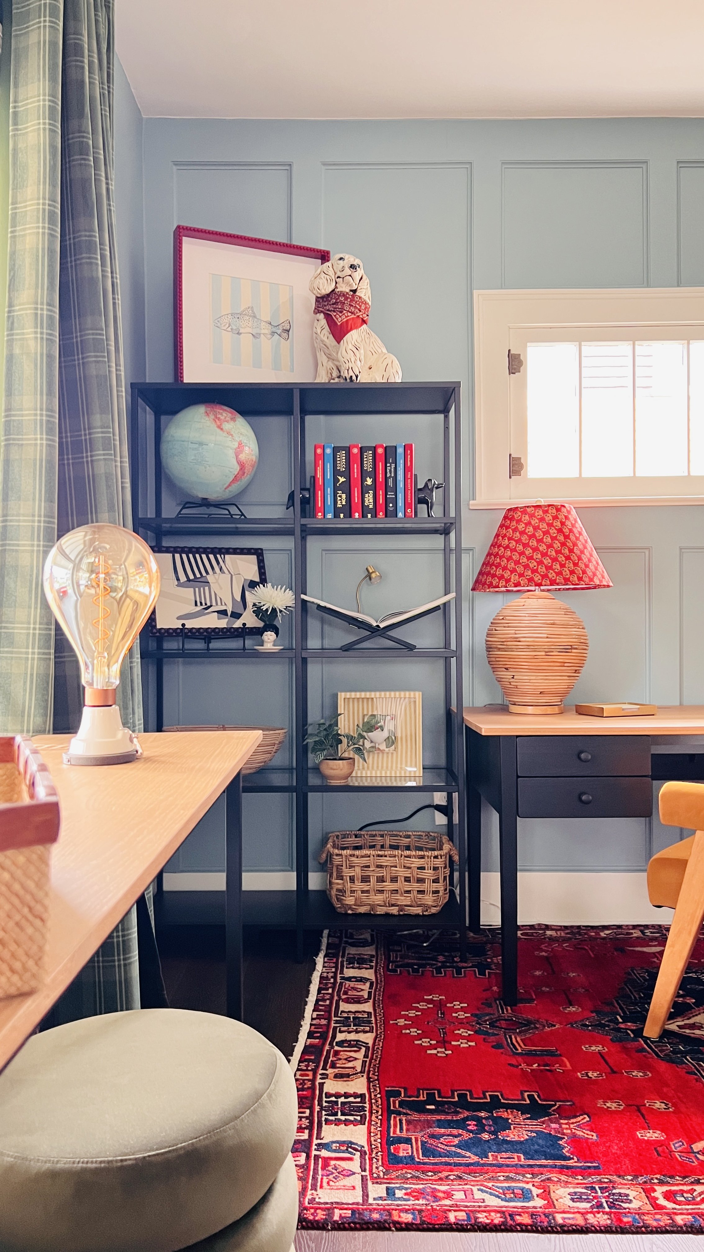 Home office with desk and shelving. Features a lamp with a red patterned shade, globe, books, decorative figurine of a dog, framed art, and a red area rug. The room has plaid curtains, a window, and wicker baskets.