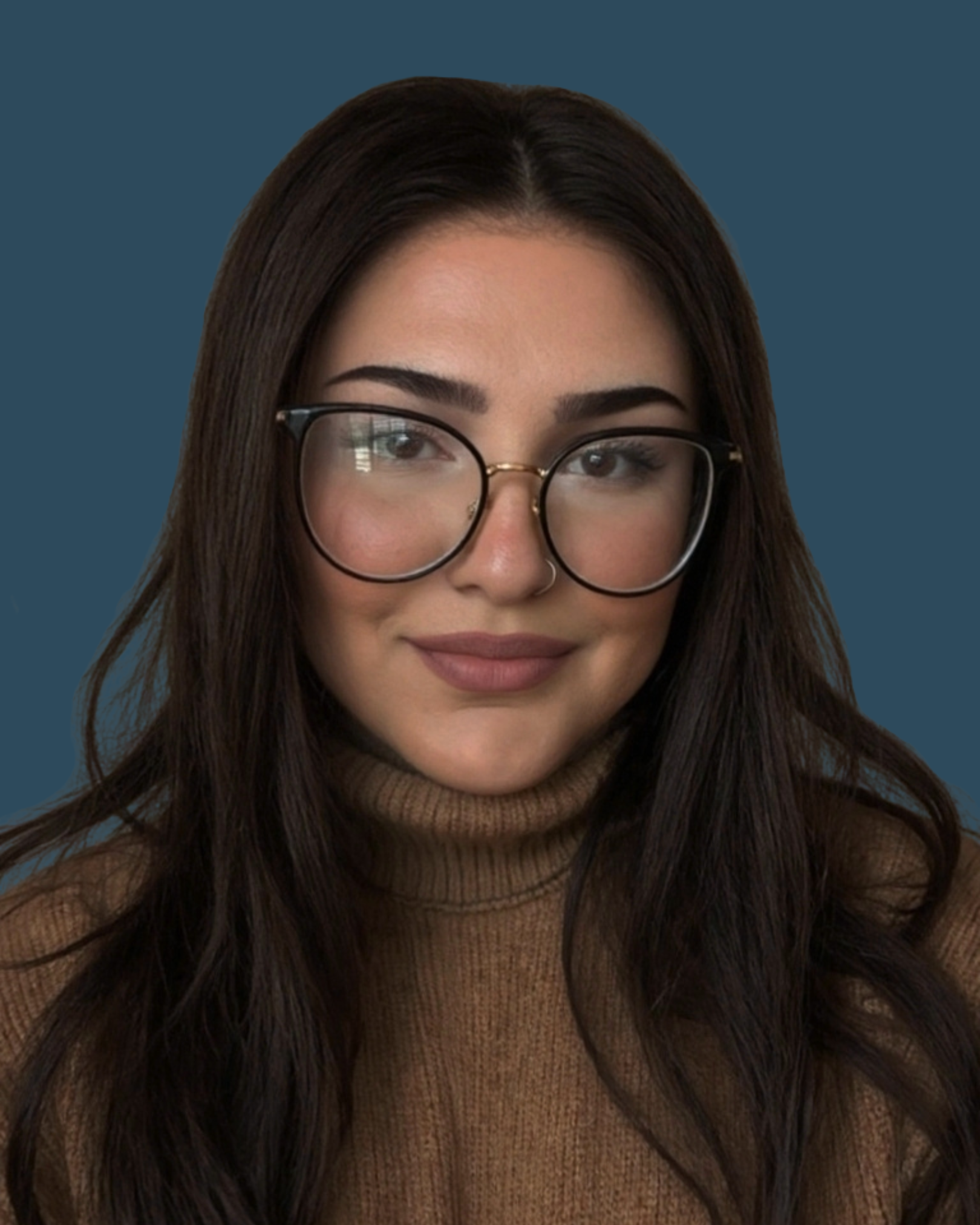 A woman with long dark hair, wearing glasses, a nose ring, and a brown turtleneck sweater, smiling in front of a dark blue background.