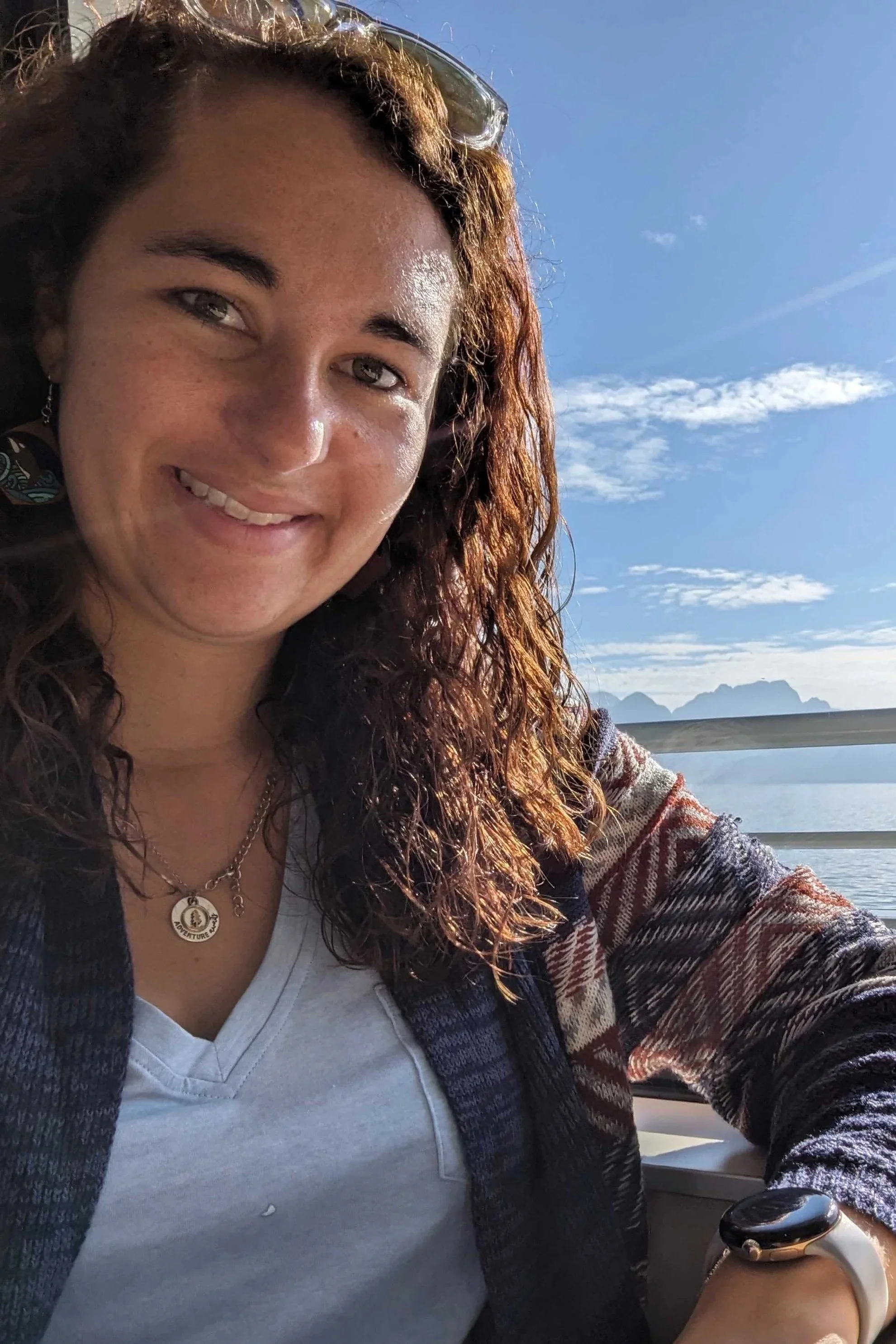 A woman with long curly hair taking a selfie on a boat or ferry. She is smiling, wearing a white shirt, layered necklaces, and a patterned cardigan. In the background, there is a large body of water, distant mountains, and a partly cloudy sky.