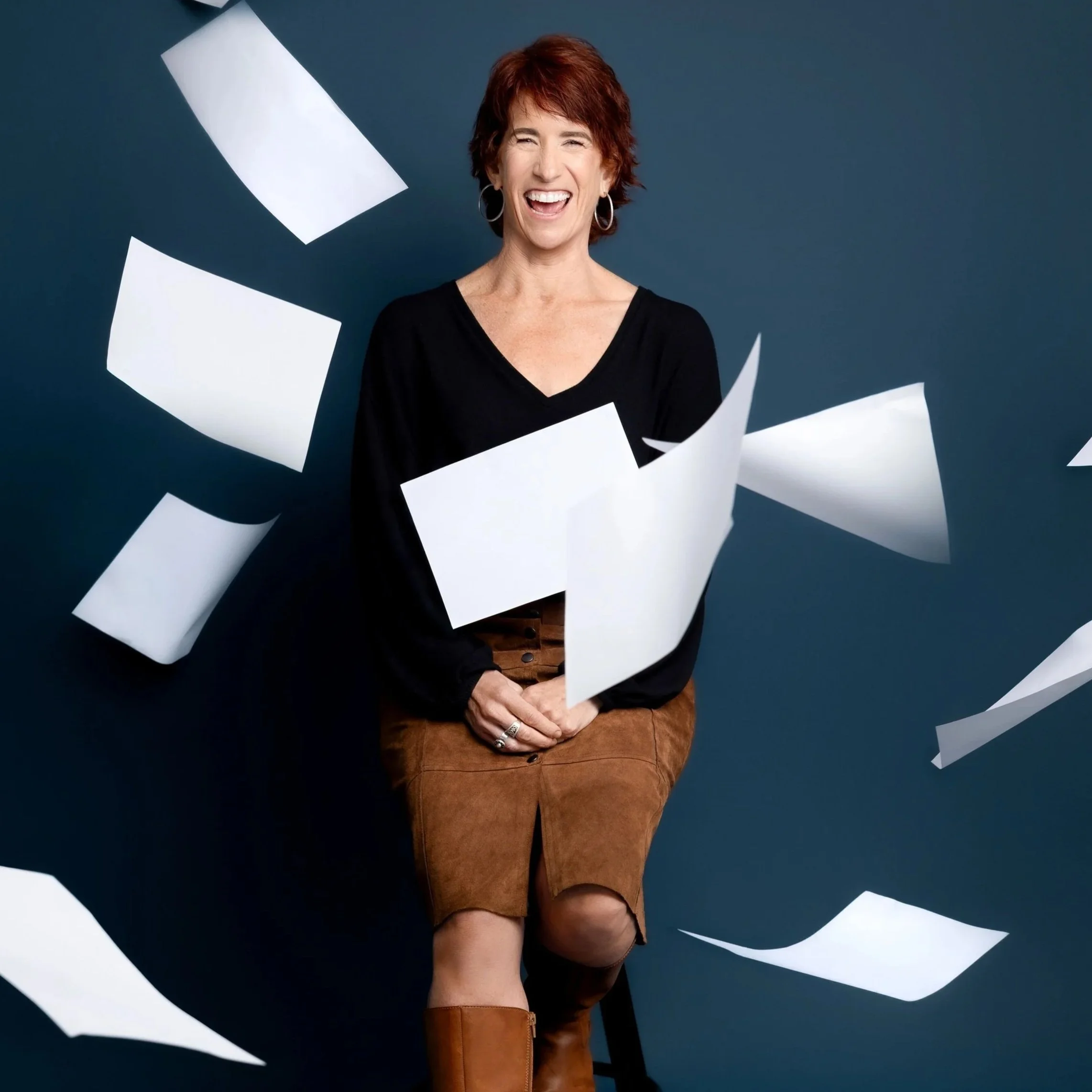 A woman with short red hair, wearing hoop earrings and a black top, smiling and posing against a dark blue background with white papers flying around her.
