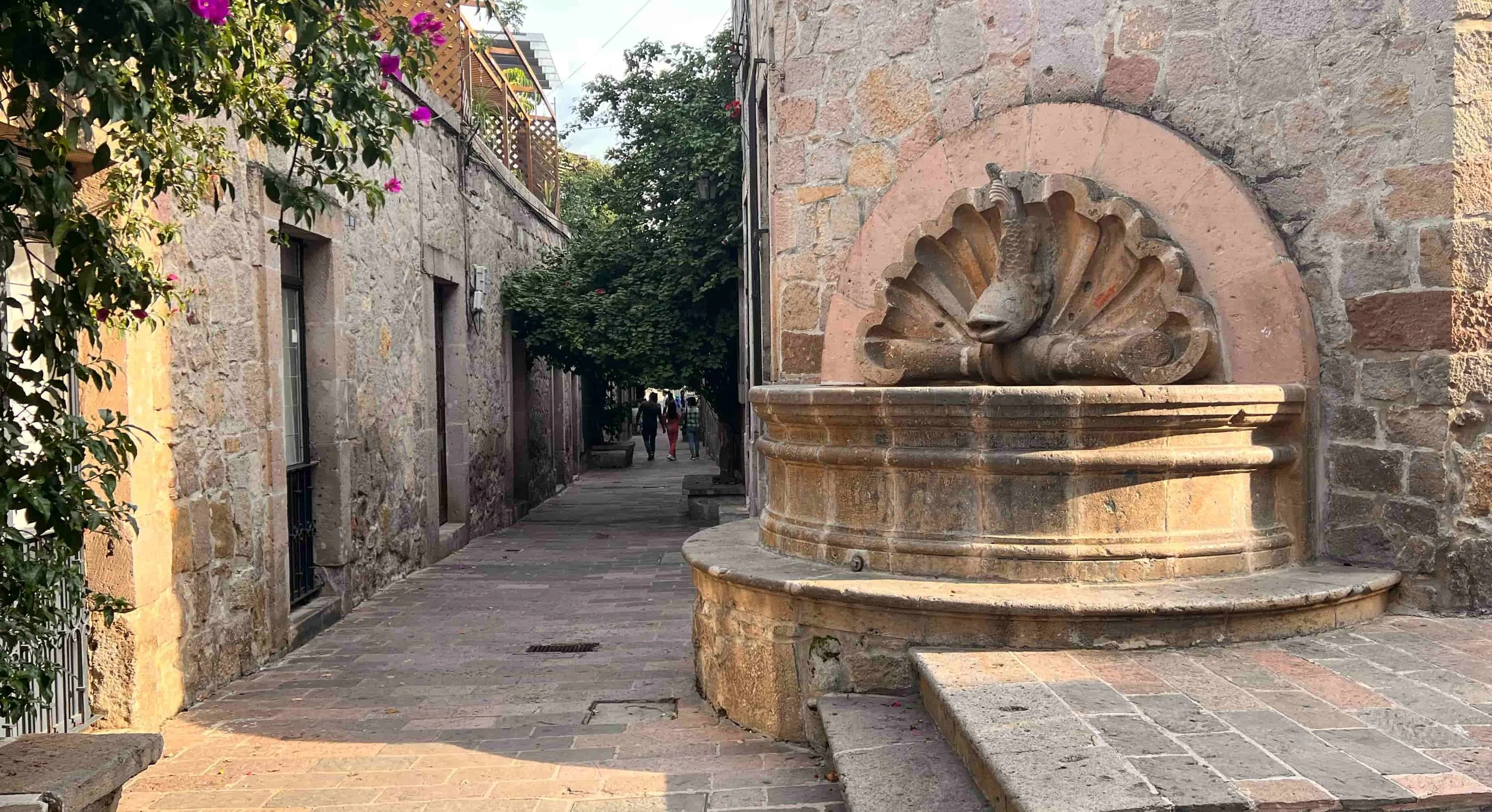 Fuentes de piedra con escultura de pavo real, en una calle empedrada con árboles y paredes de piedra, en un pueblo antiguo.