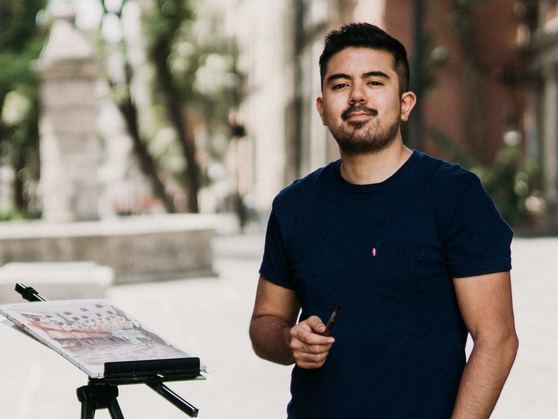 Joven pintor con barba y camiseta azul, sosteniendo un pincel, en una calle con caballete y pintura.
