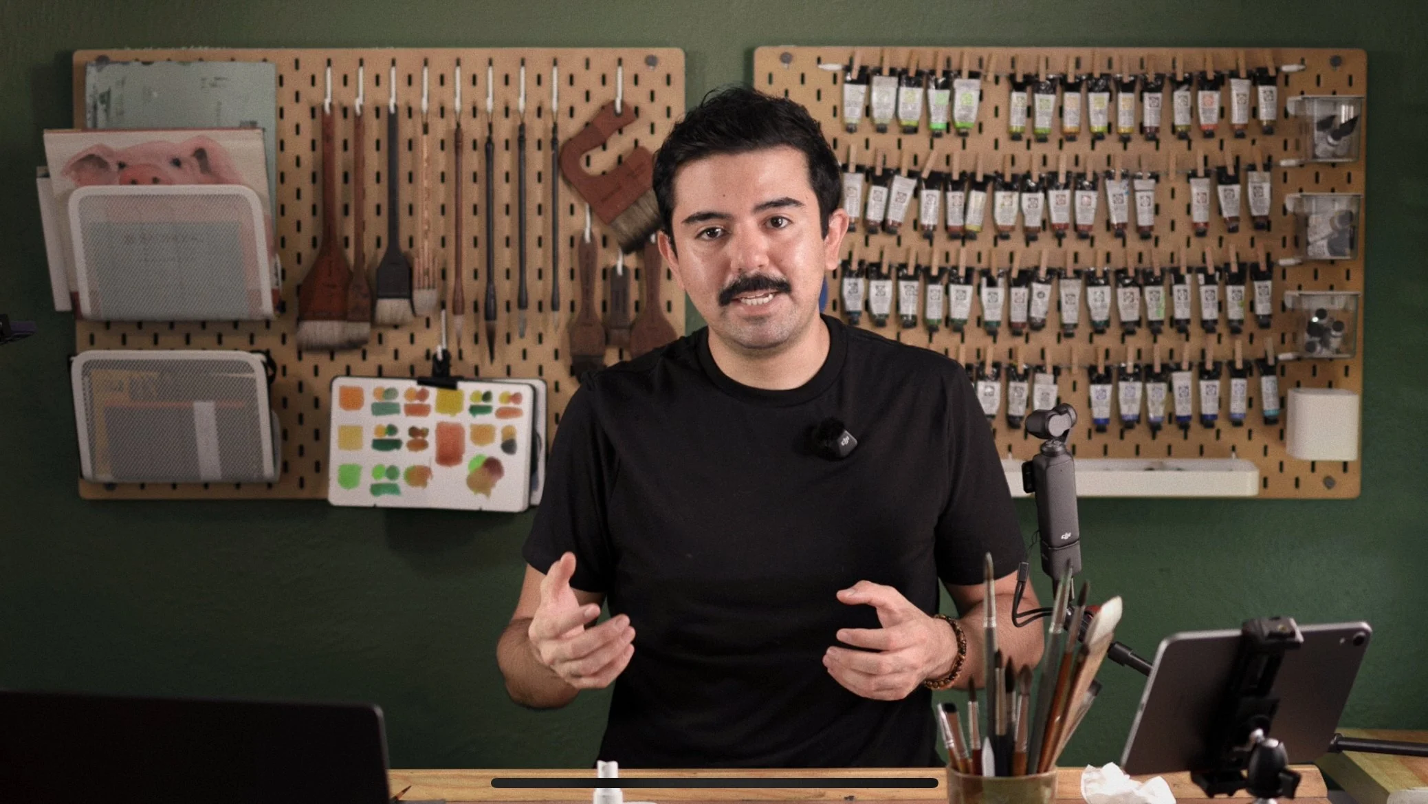 Hombre con bigote y camiseta negra, sentado en un taller con herramientas y pinturas en una pared de fondo, hablando y gesticulando.