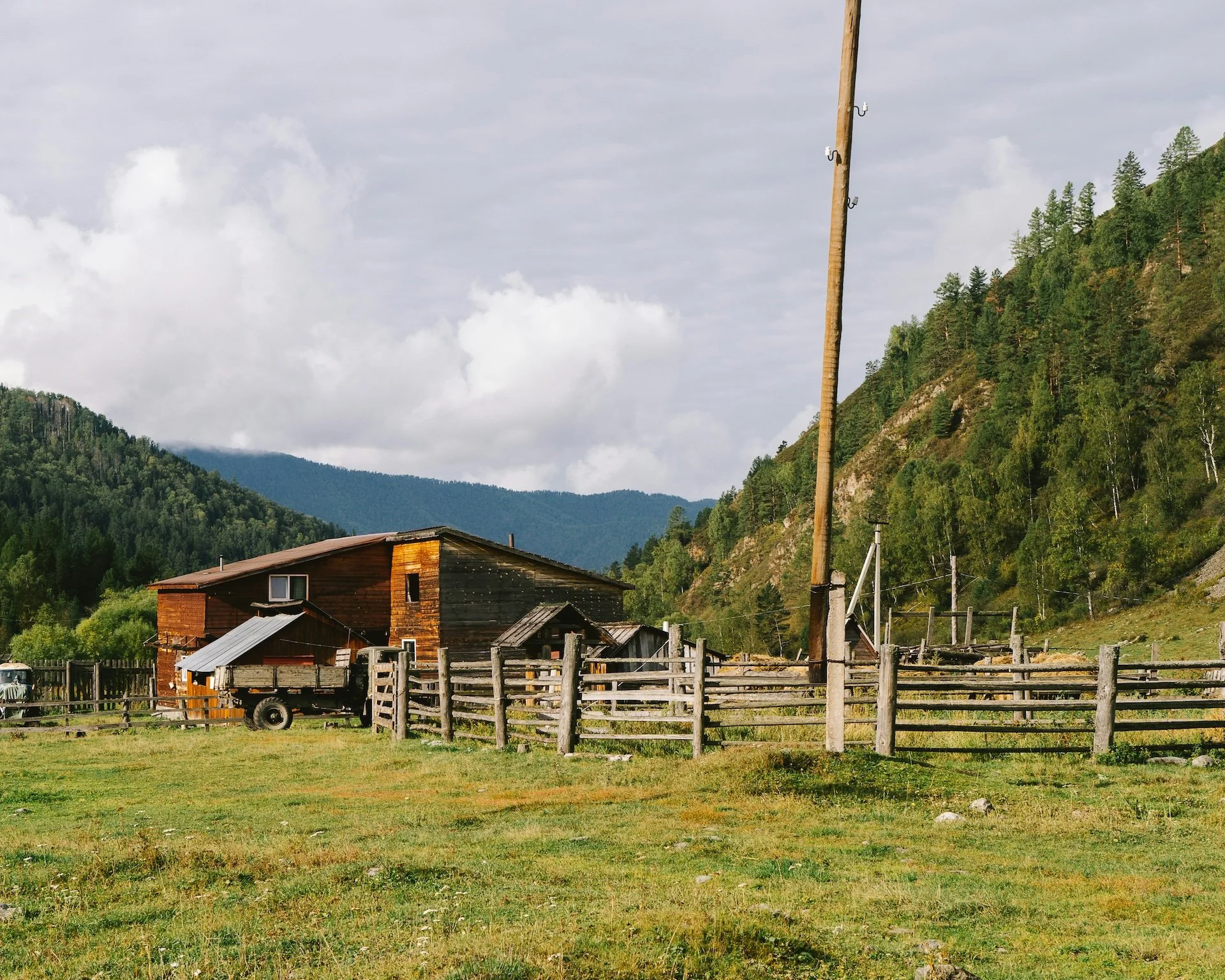 Rural landscape with a wooden barn, fences, and a wagon, surrounded by hills and trees under a cloudy sky.