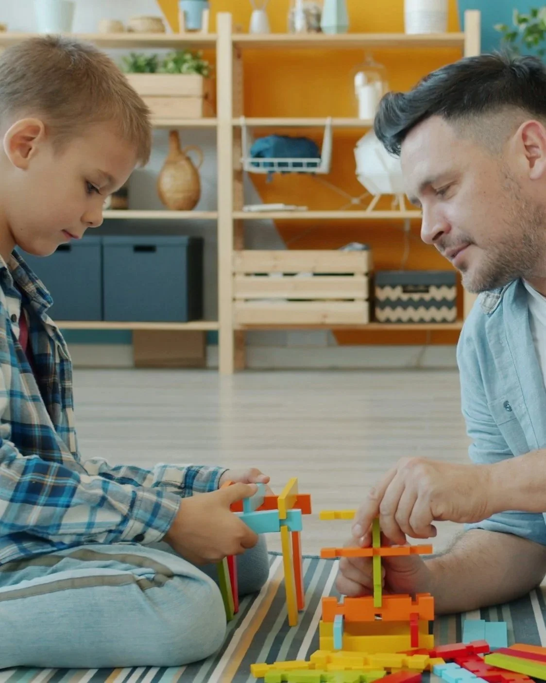 A young boy and an adult man playing with colorful building blocks on a striped surface, in a bright room with shelves and decorations in the background.