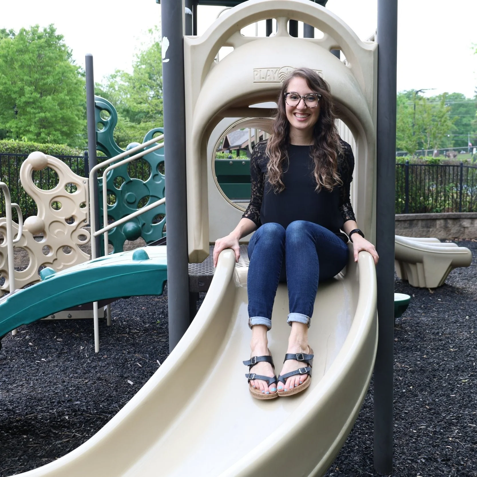 A young woman with glasses, curly hair, and a black lace top sitting at the top of a slide at a playground, smiling.