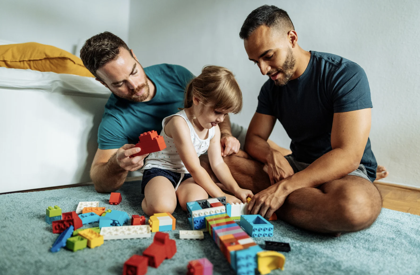 Two men and a young girl sitting on the floor playing with colorful LEGO bricks.