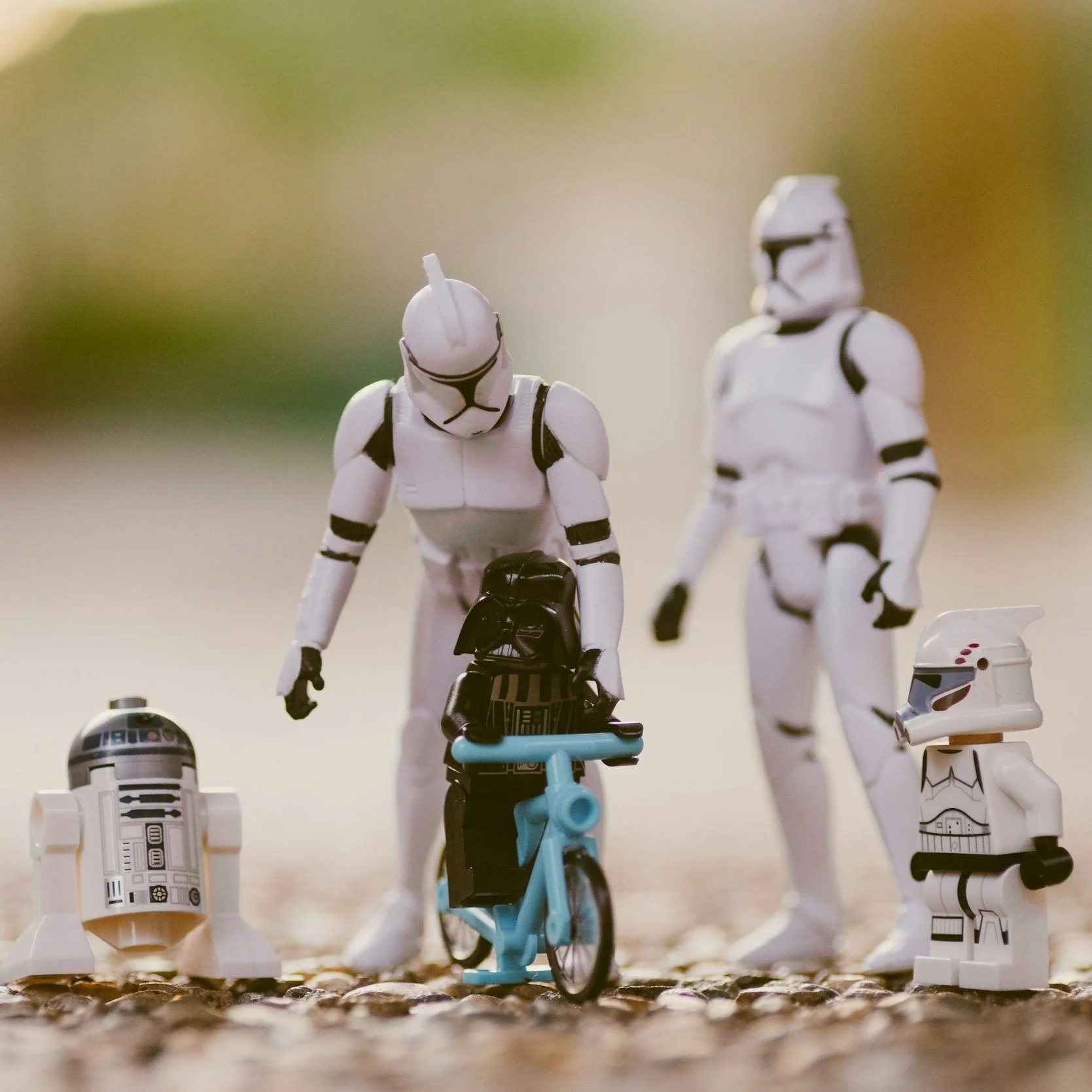 Star Wars toy figures including R2-D2, a stormtrooper, Darth Vader, and a toddler-sized stormtrooper with a small bicycle on a textured surface.